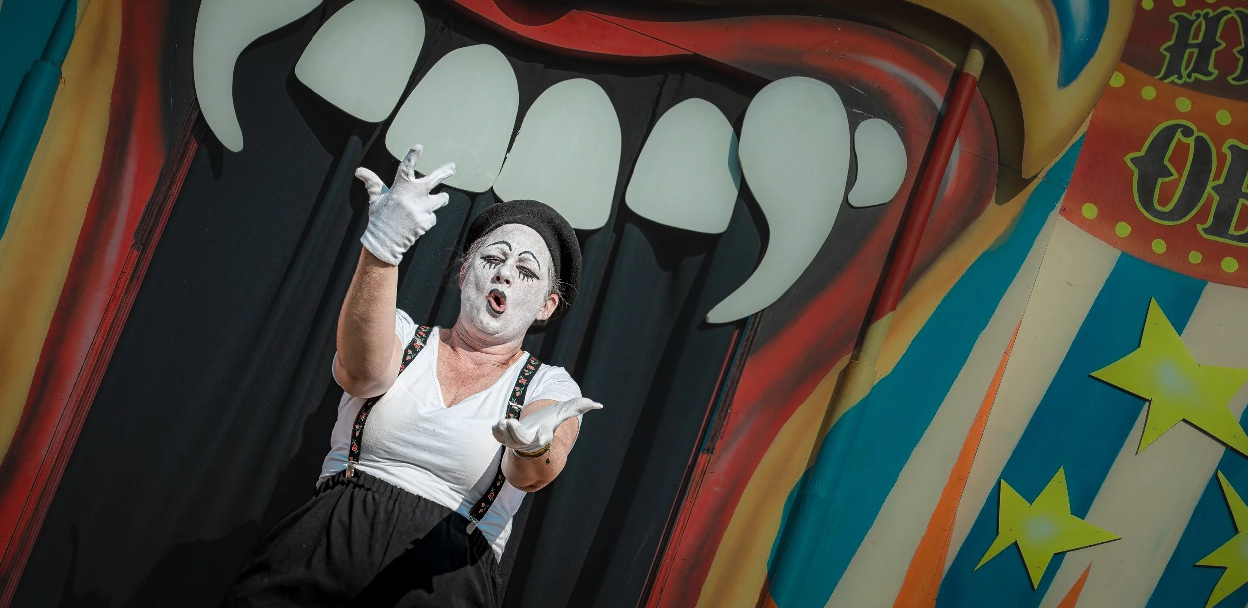 Een clown met wit gezichtsmake-up, een zwarte hoed, een witte shirt en zwarte broek die danst op een circuspodium met kleurrijke decoraties en een grote mond met tanden op de achtergrond.