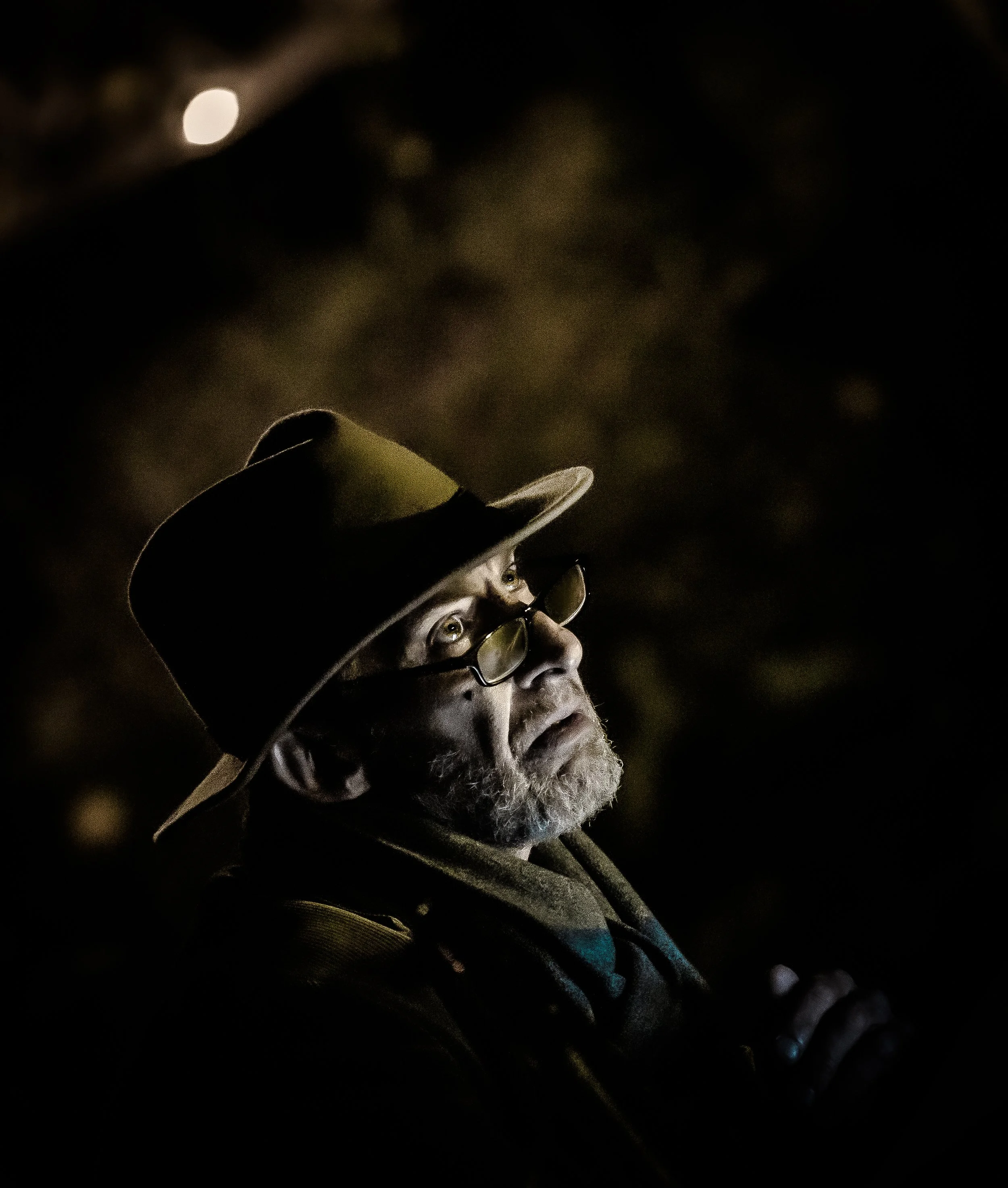 Een oudere man met een hoed en bril kijkt omhoog in de donkere nacht, met een achtergrond die niet duidelijk zichtbaar is.