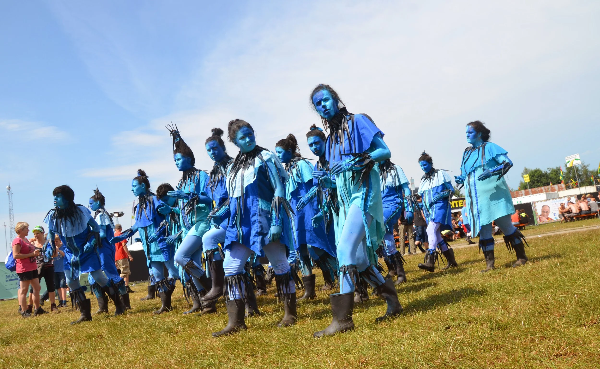 Een groep mensen in blauwe kostuums met blauwe make-up en zwarte accessoires tijdens een outdoor festival.