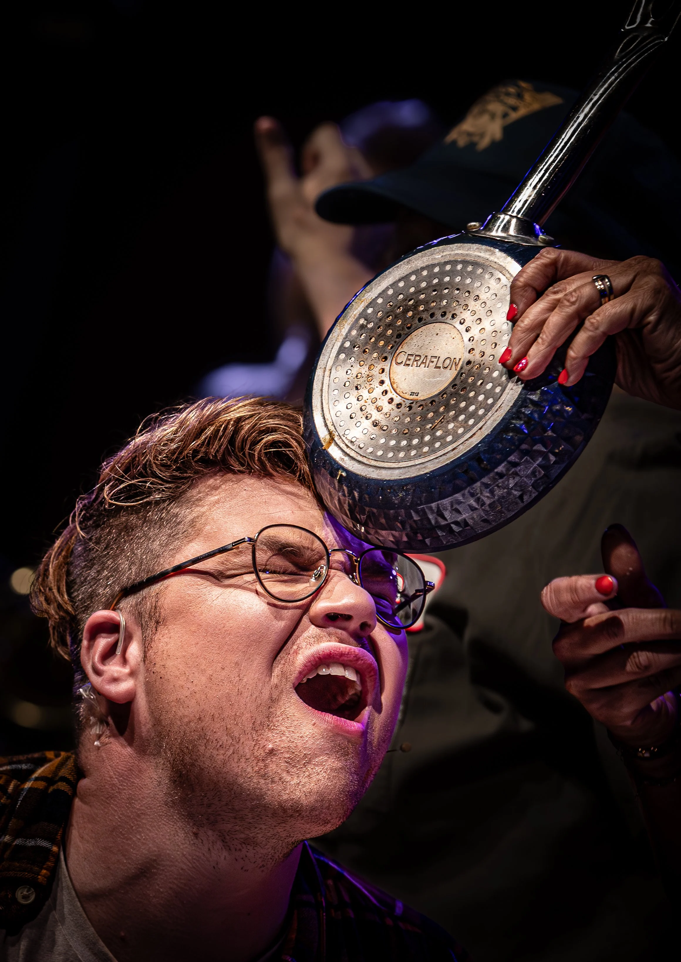 Een man met een bril die zich pijn doet en ergert terwijl er een pan wordt geslagen op zijn hoofd door een ander persoon. De man heeft een open mond en lijkt te schreeuwen.