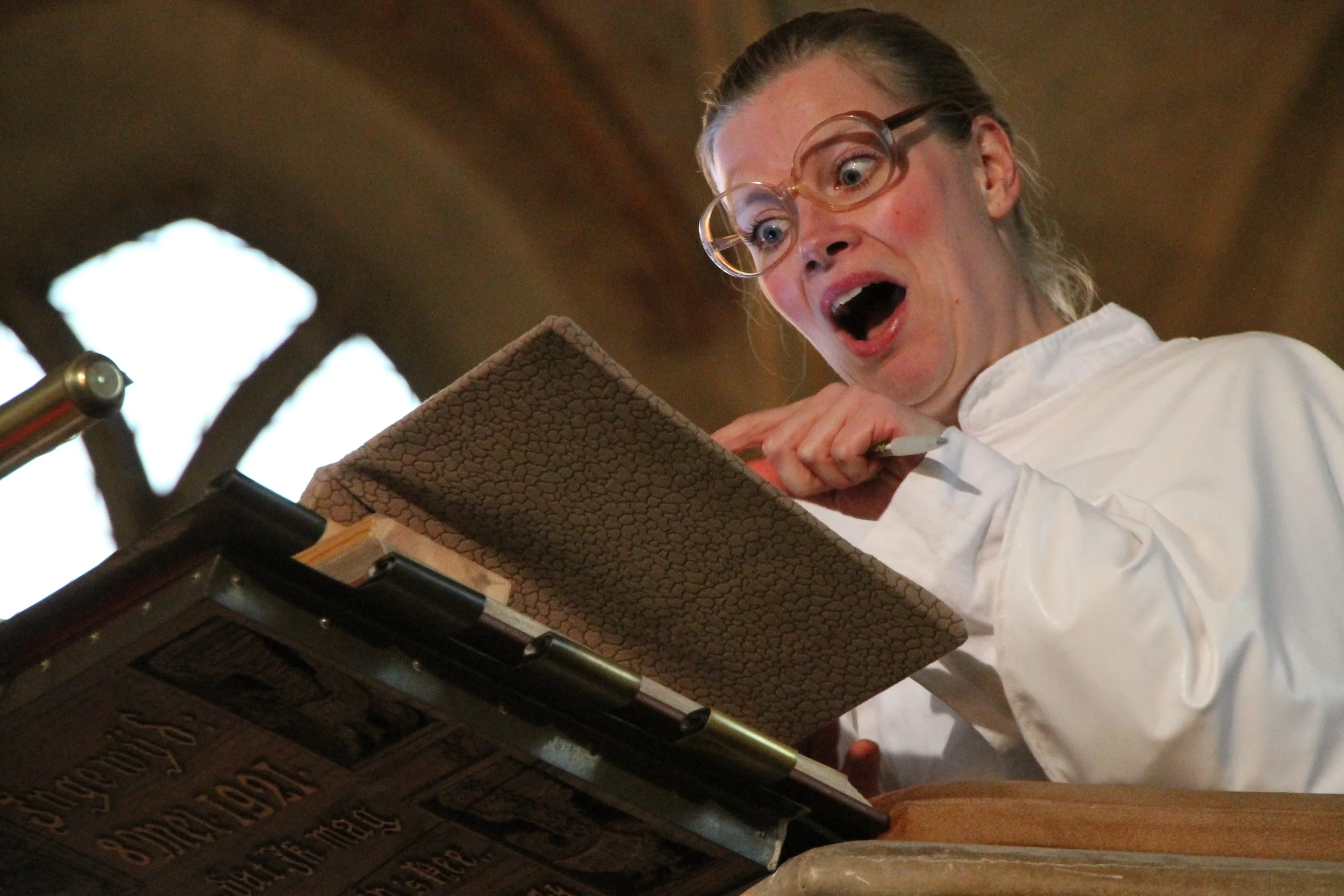 Vrouw met bril en witte jas kijkt verbaasd naar een boek of script op een lessenaar in een kerk of oude zaal.