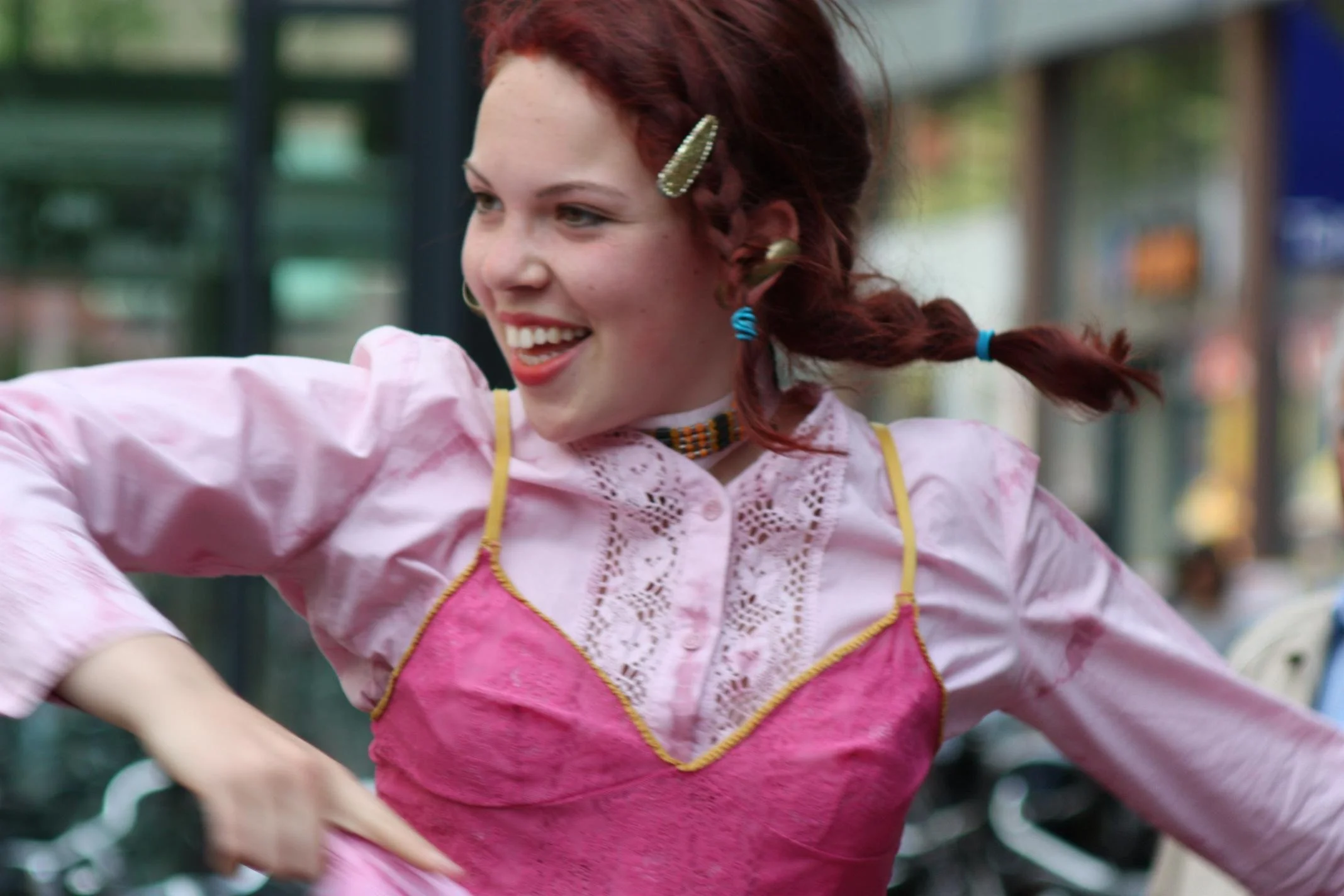 Vrouw met rood haar in een gevlochten kapsel met haarspelden, lachend in een roze jurk met gele en blauwe details, op een straat met fietsen op de achtergrond.