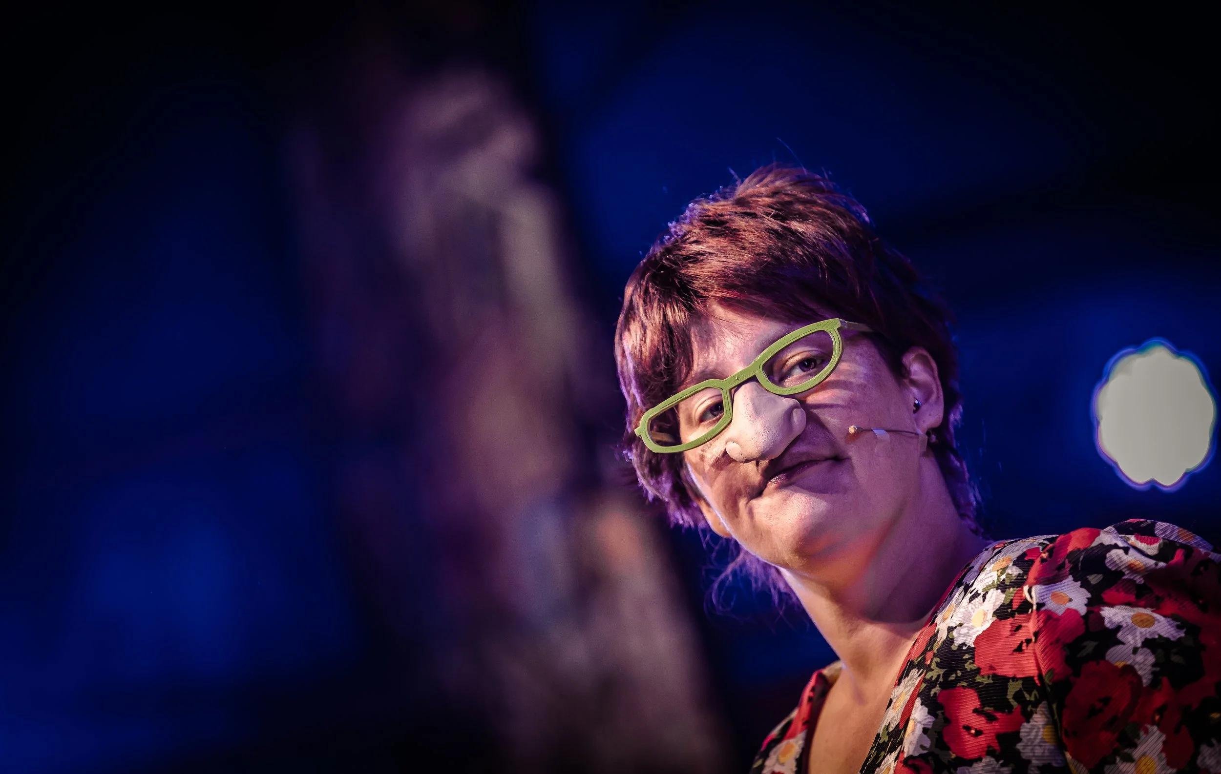 Een vrouw met groene brillen en een lach, op een podium met blauwe achtergrondverlichting.