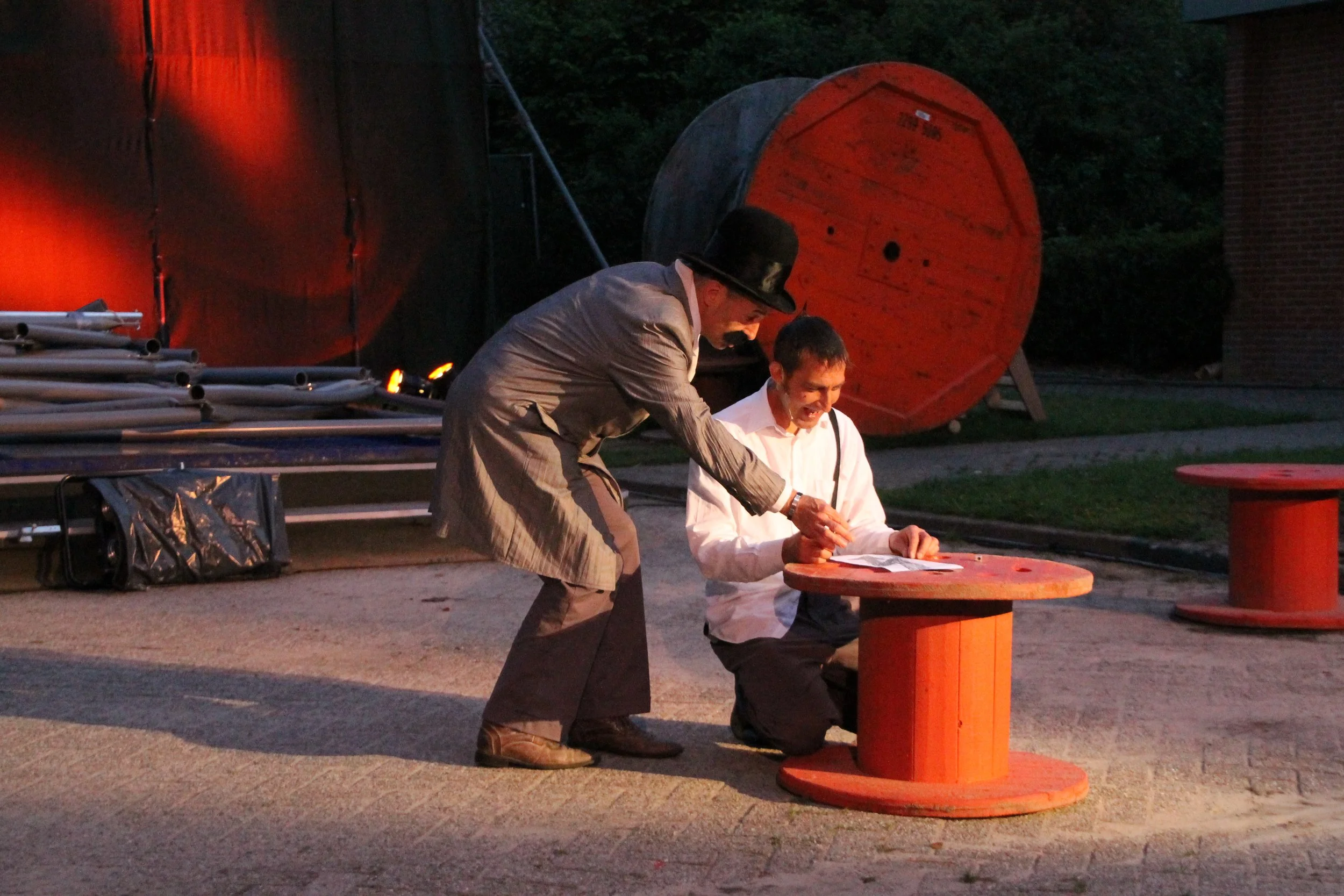 Twee mannen in vintage kleding, een staat wijzend terwijl de ander schrijft, op een roodgeverfd houten tafel buiten bij schemerlicht.