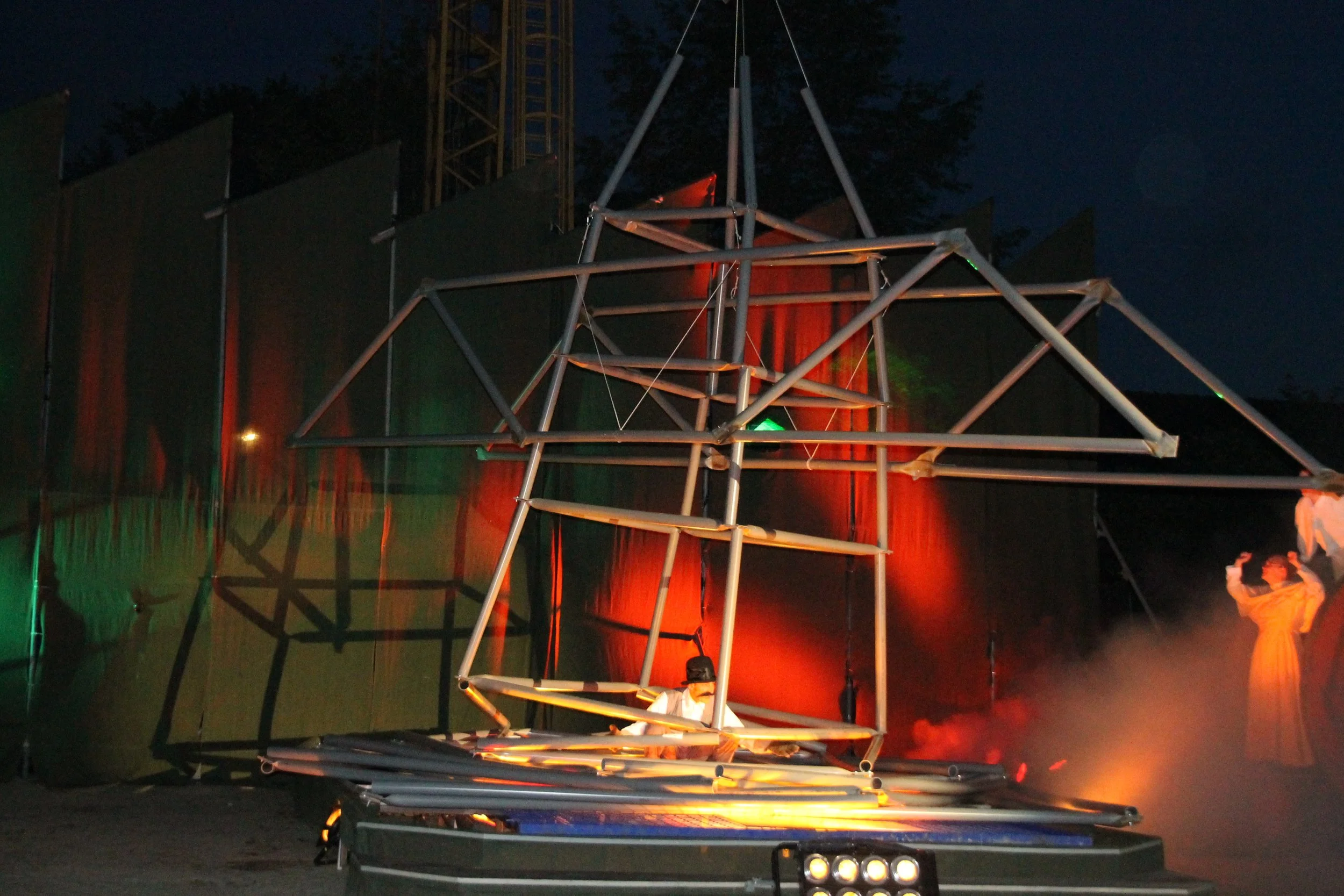 Een metalen structuur die lijkt op een geometrisch doolhof, met kleurrijke achtergrondverlichting, tijdens de avond of nacht.