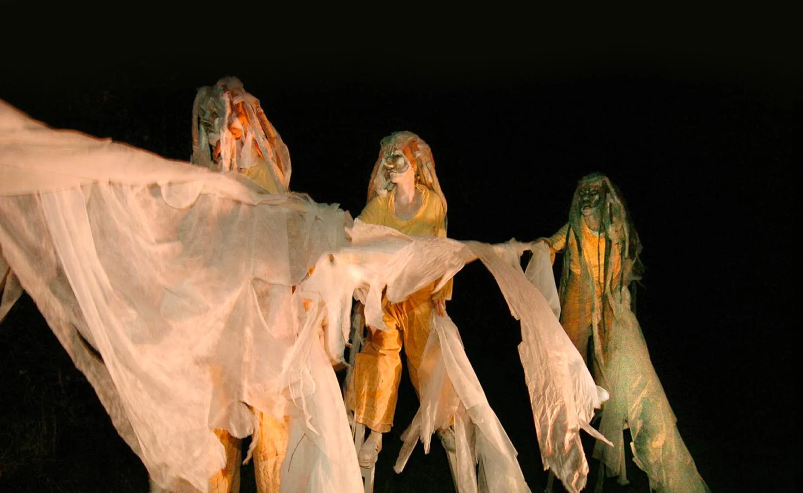 Drie personen gekleed in kleurrijke, abstracte stoffen kleden die lijken op performance kunst, tegen een zwarte achtergrond.