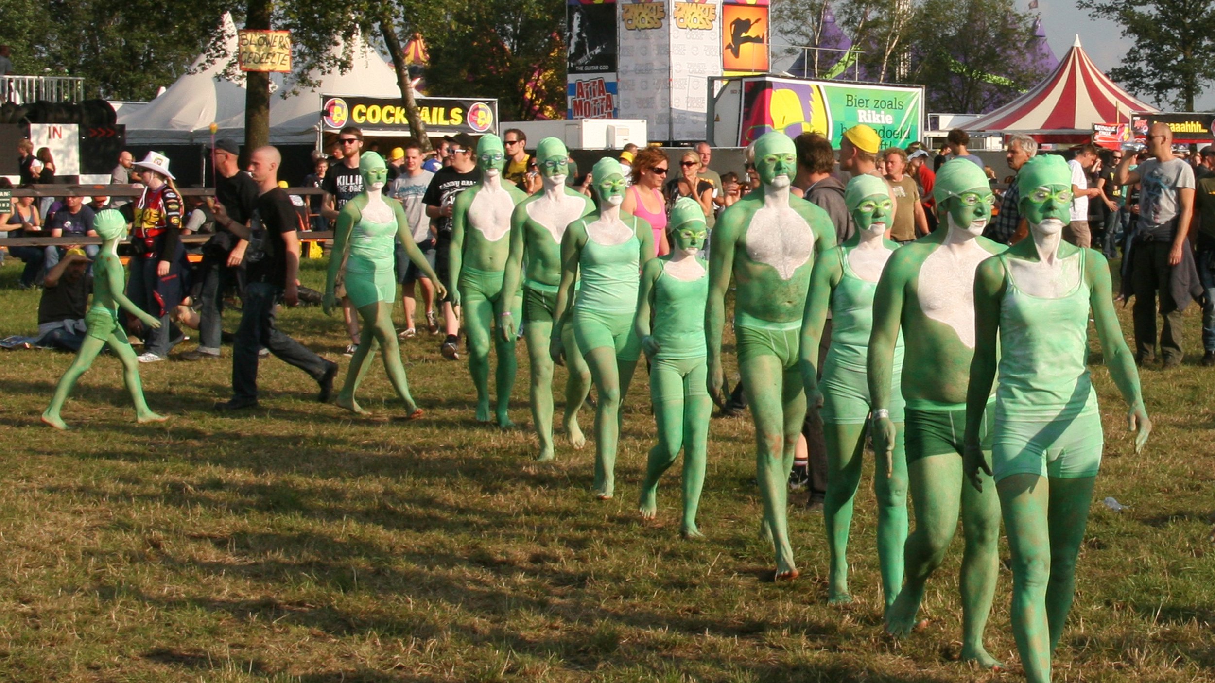 Verschillende mensen in groene lichaamsverf en kostuums lopen over een festivalweide, met eet- en drankstandjes en een groot scherm op de achtergrond.