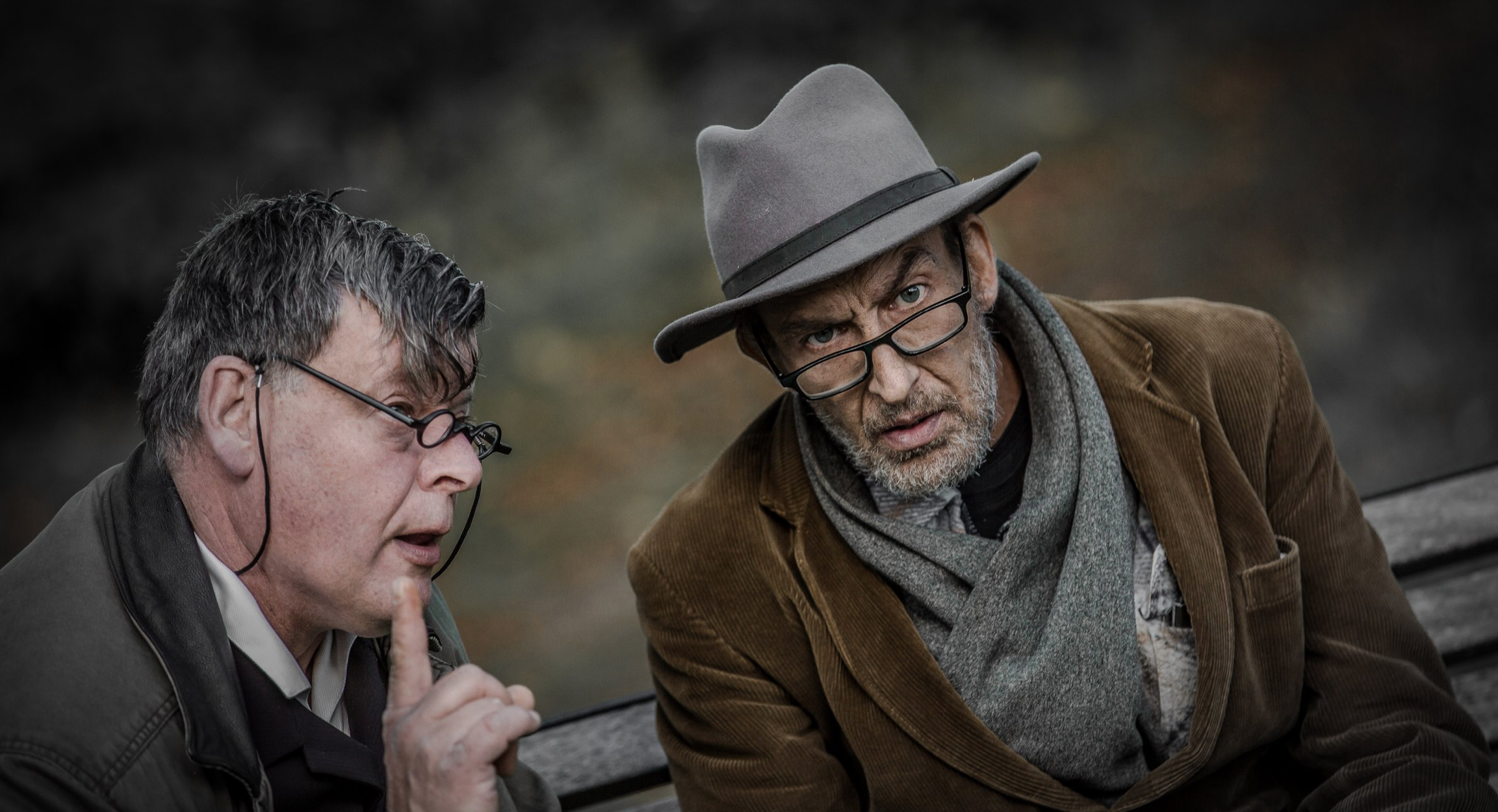 Twee oudere mannen met brillen zitten op een bank, de ene man draagt een hoed en kijkt serieus terwijl de andere man zijn vinger omhoog houdt in een stille gebaar, op een bank in een park of natuurlijke omgeving.
