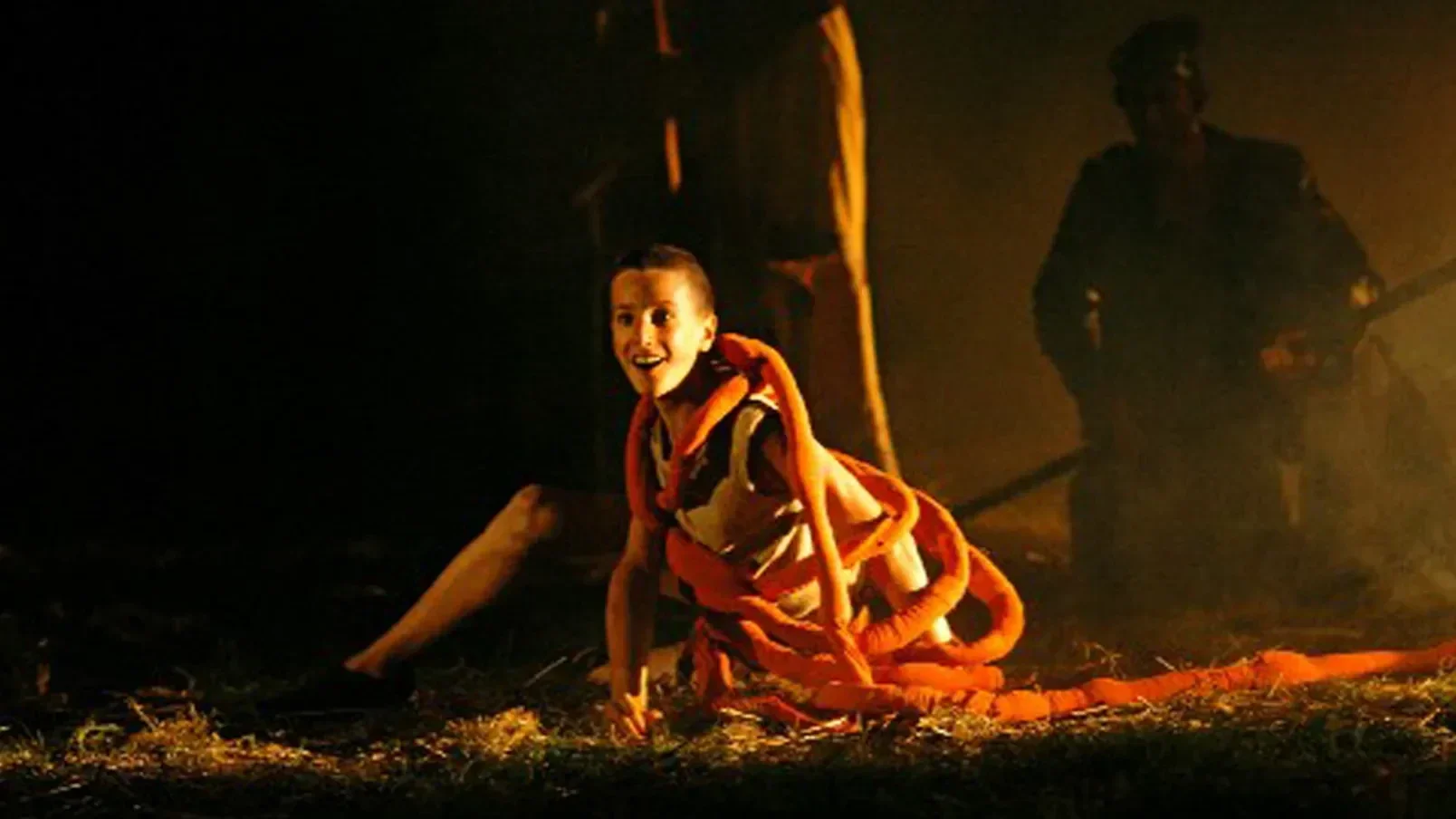 Een persoon op de grond tijdens een toneelstuk, met een persoon op de achtergrond met een gitaar, in een donkere setting met oranje verlichting.