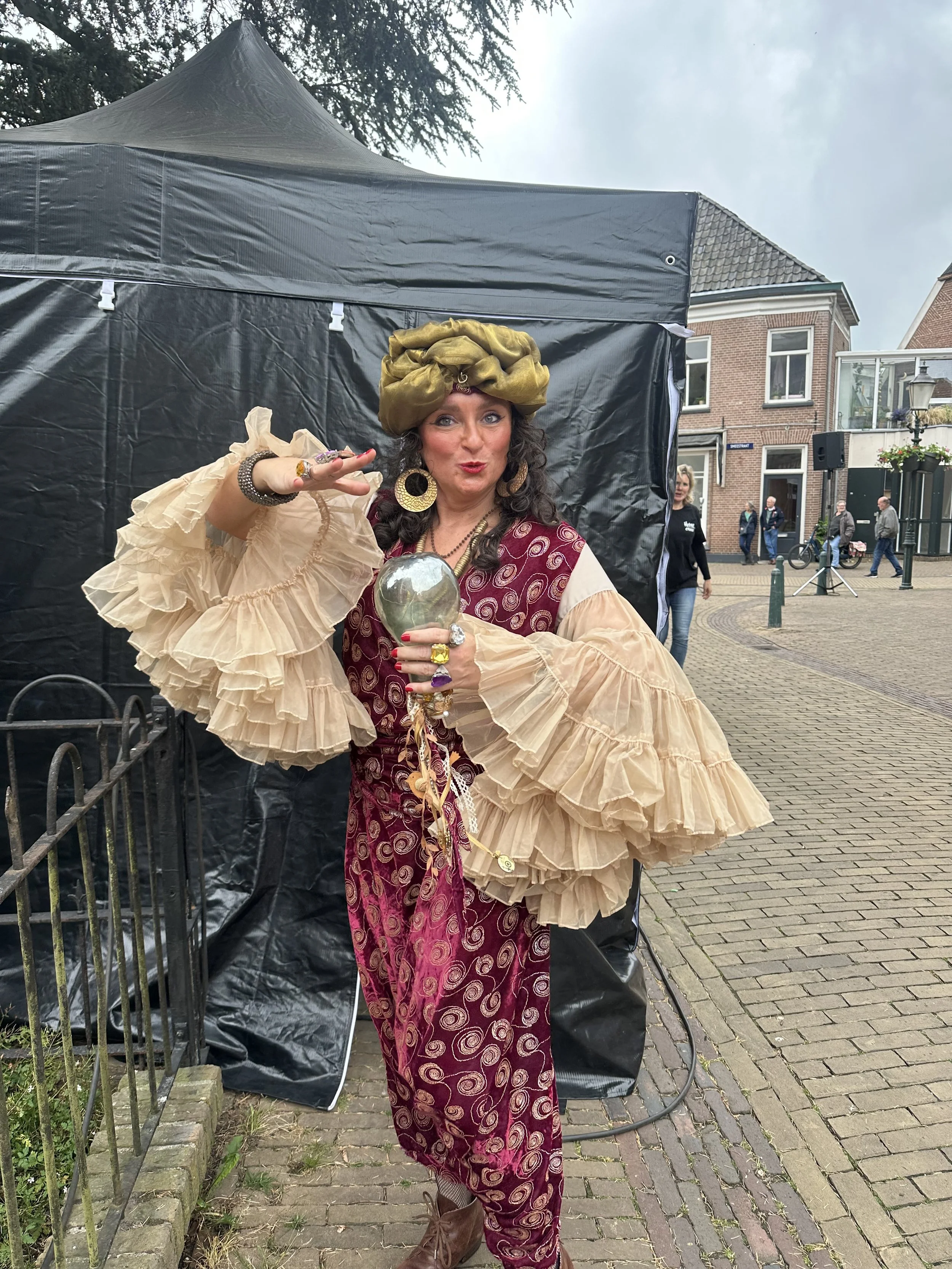 Een vrouw in een kleurrijk kostuum met grote ruffles en sieraden poseert voor een zwart tentdoek op een straat met mensen op de achtergrond.