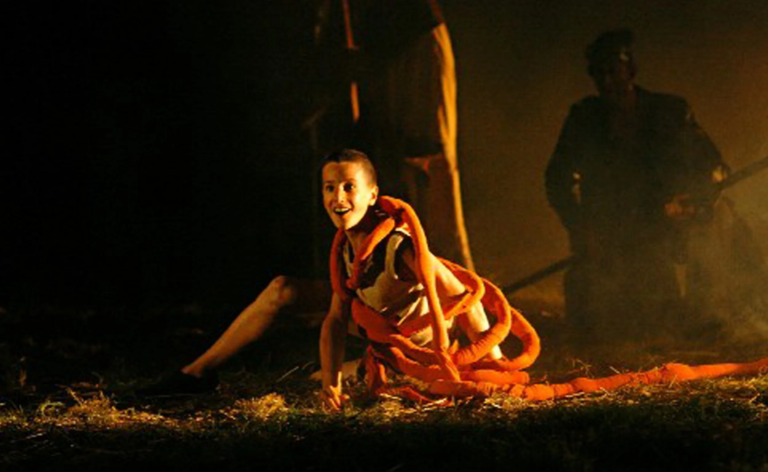 Kind met een lach op zijn gezicht die op de grond ligt, gekleed in een kostuum van oranje gekrulde tentakels, met een donkere achtergrond.