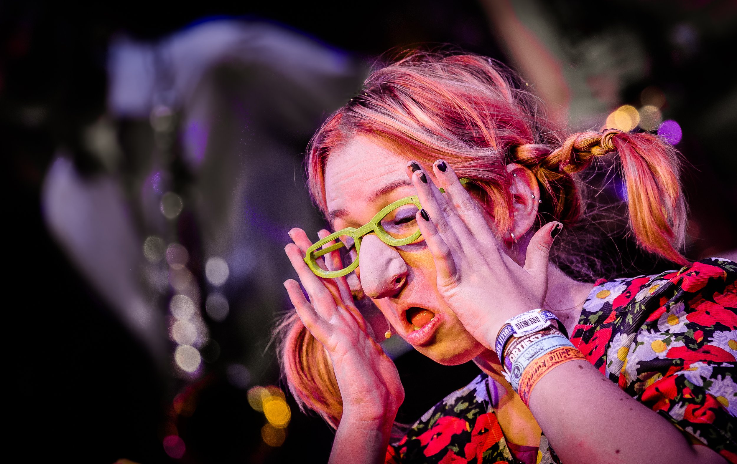 Vrouw met roze haar, groene bril, verklaart met handgebaren, kleurrijk shirt, op een levende muziekfestivalevenement met achtergrondlichten.