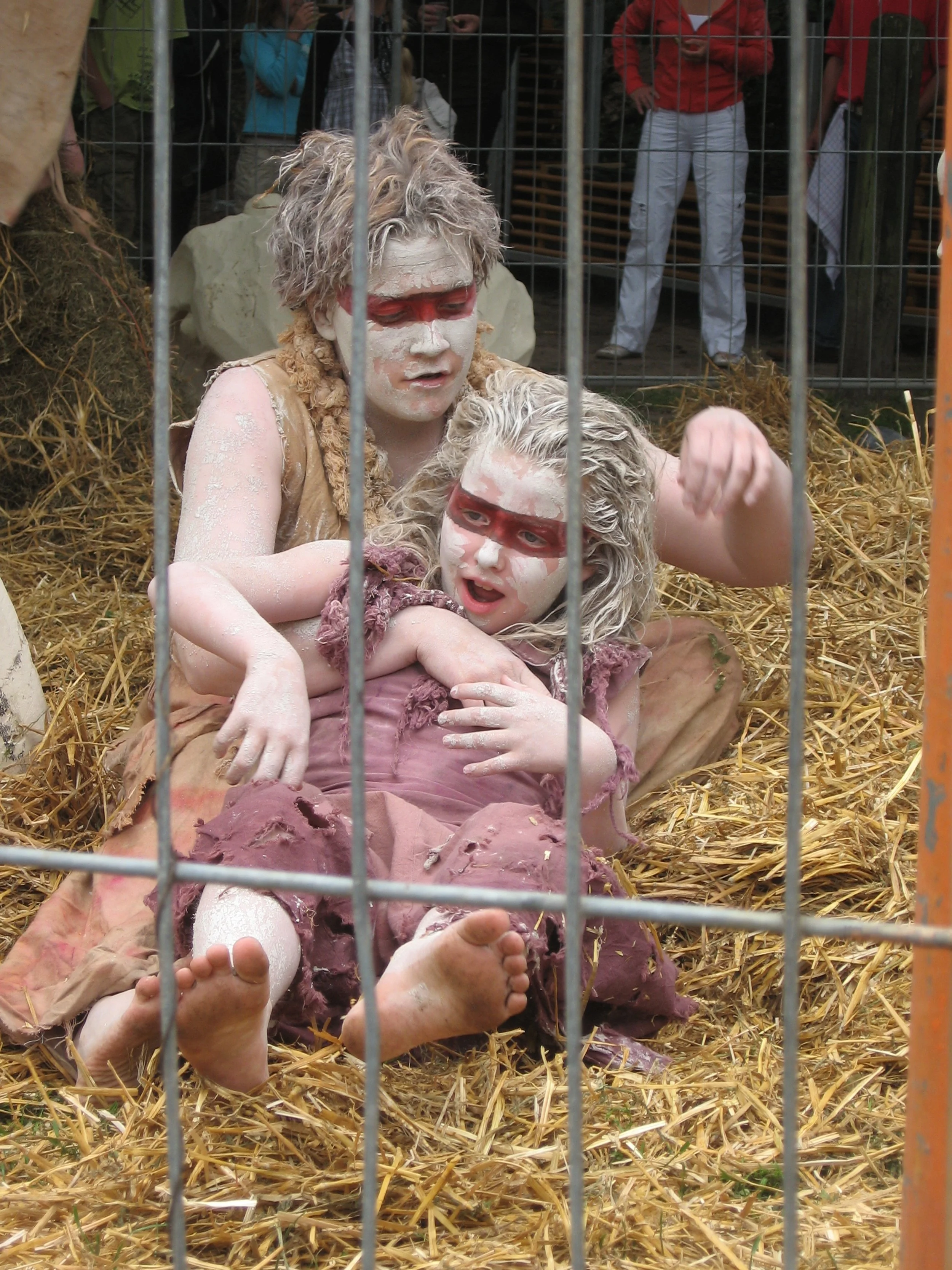 Twee mensen verkleed als straatartiesten, met witte schmink en rode aangezichten, zitten op stro. Ze lijken te doen alsof ze in een kooi zitten, met een metalen hek ervoor. Ze zijn in een buitenomgeving, omringd door bezoekers.