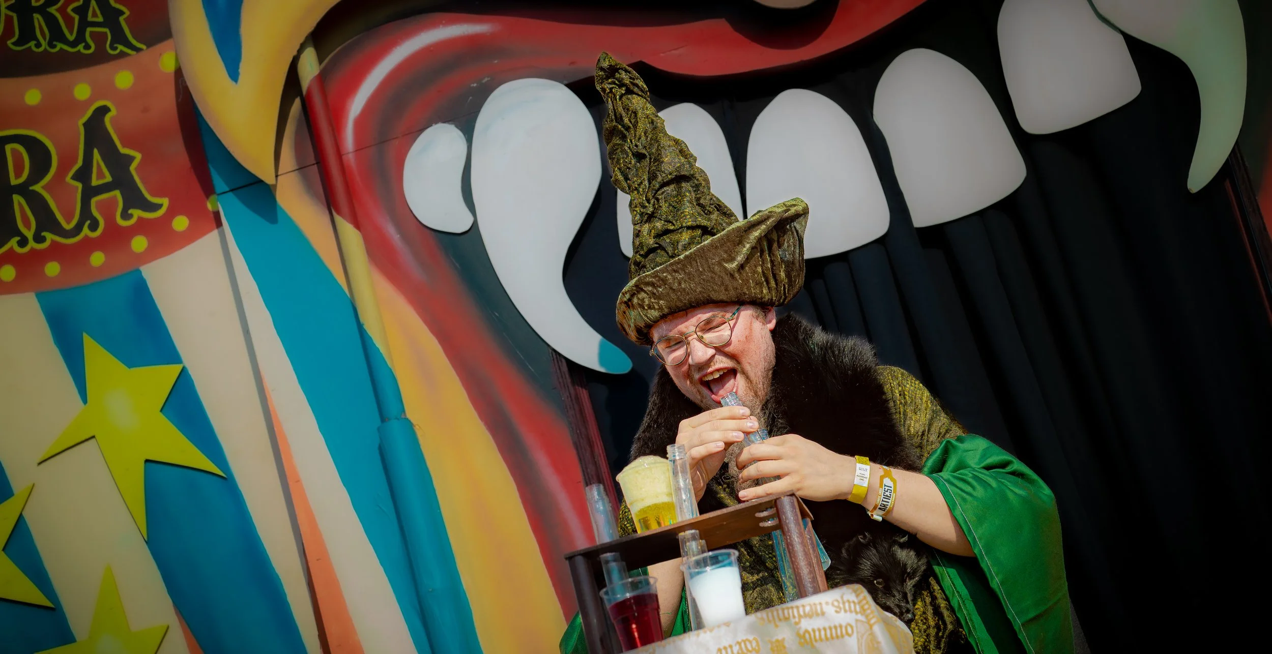 Man in fantasierijk kostuum met een grote puntmuts, lachend terwijl hij een proefbuisje mixt, met een kleurrijk circusachtig decor en een groot opgezette mond met witte tanden op de achtergrond.