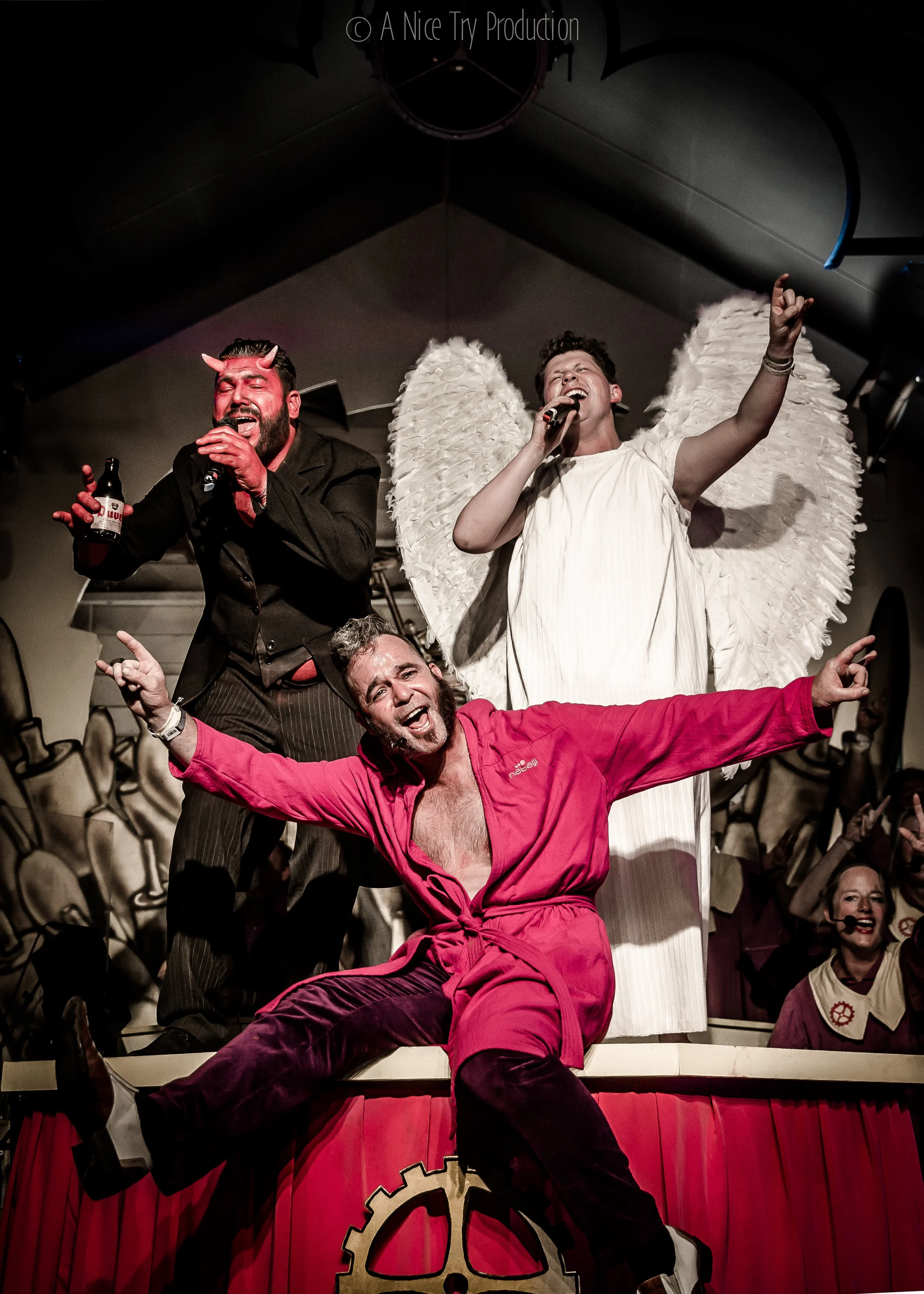 Drie mensen op een podium tijdens een theatrale uitvoering. Eén man met hoorns en een waterflesje, één persoon met grote witte vleugels en een wit jurk, en een man in een roze outfit die in een dynamische pose staat. Op de achtergrond zijn mensen te zien en decoraties.