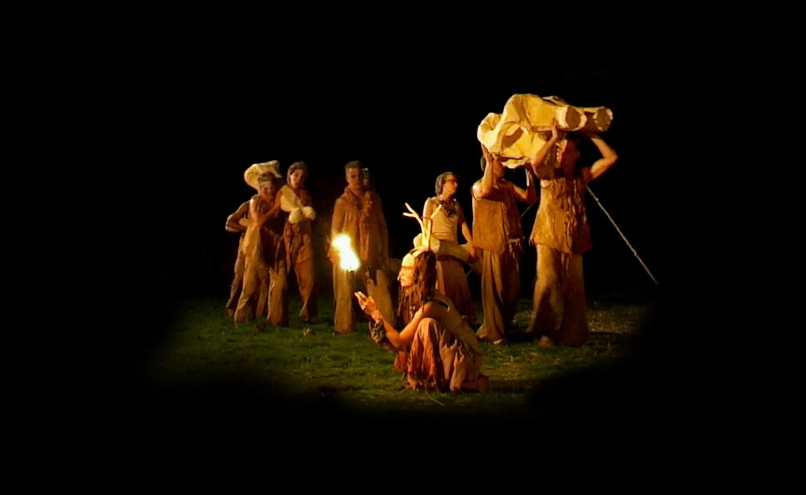 Groep mensen in kostuums tijdens een theatervoorstelling in de duisternis, met één persoon met een fakkel.