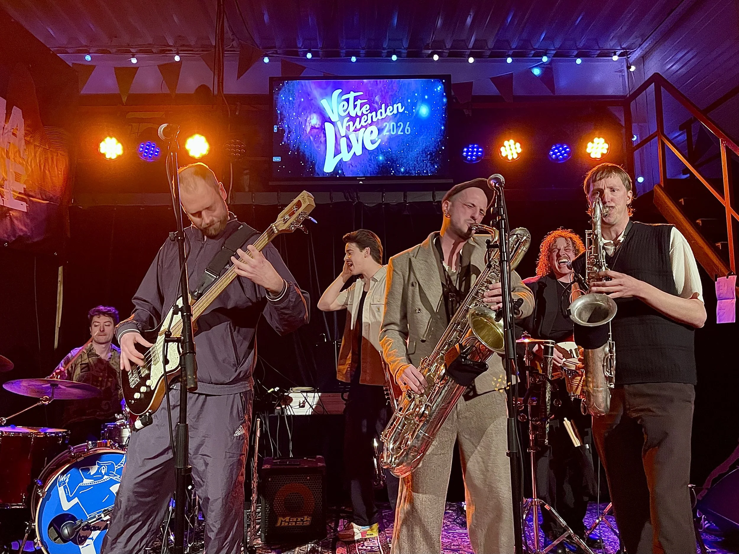 Band de Baron voert live op tijdens een optreden met kleurrijke achtergrondverlichting en een grote tv-scherm met de tekst 'Vette Vrienden Live 2026' boven hen.