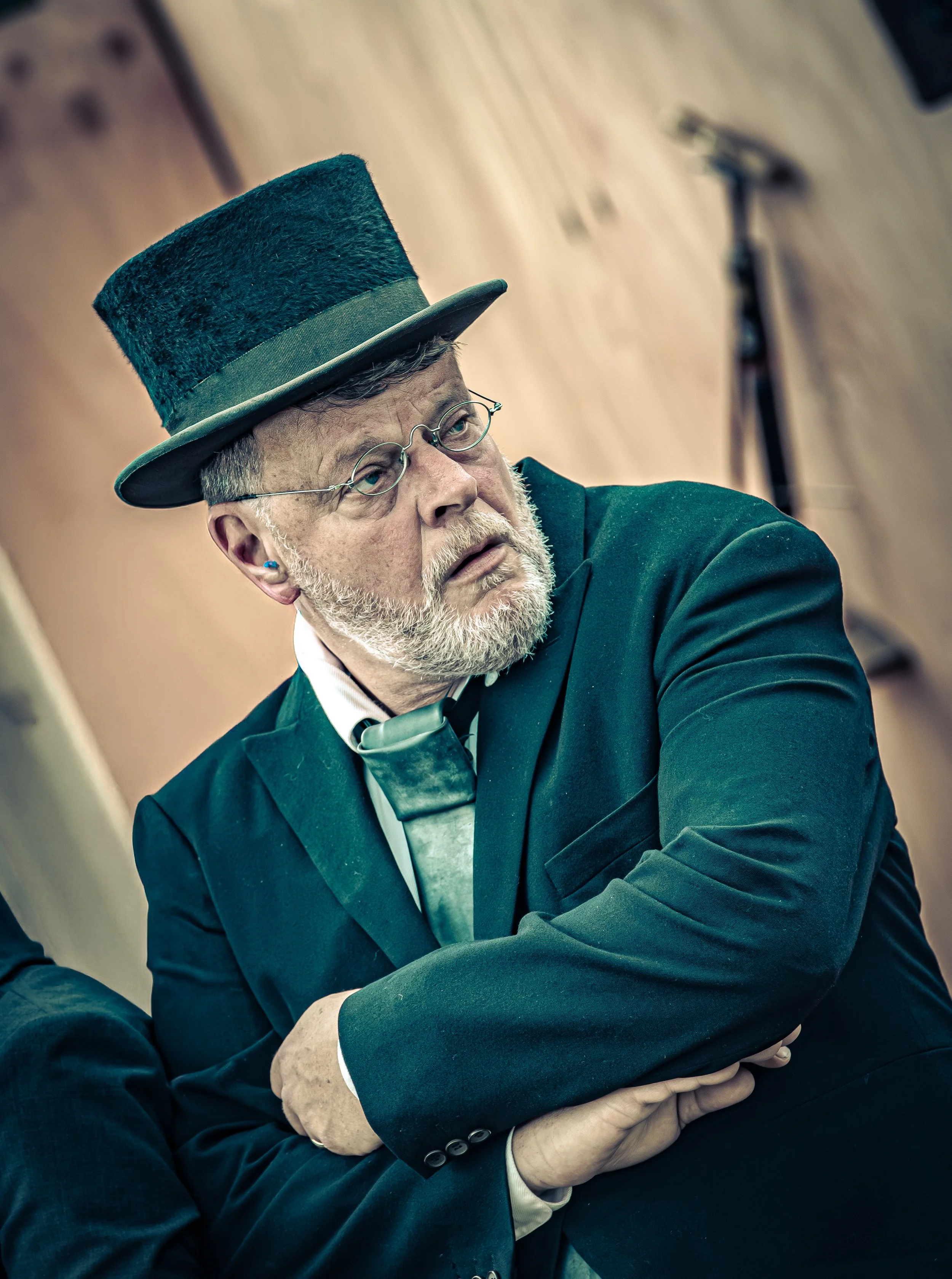 Man in historische kostuum en hoge hoed, met bril en grijs haar, in een gesprek of presentatie.