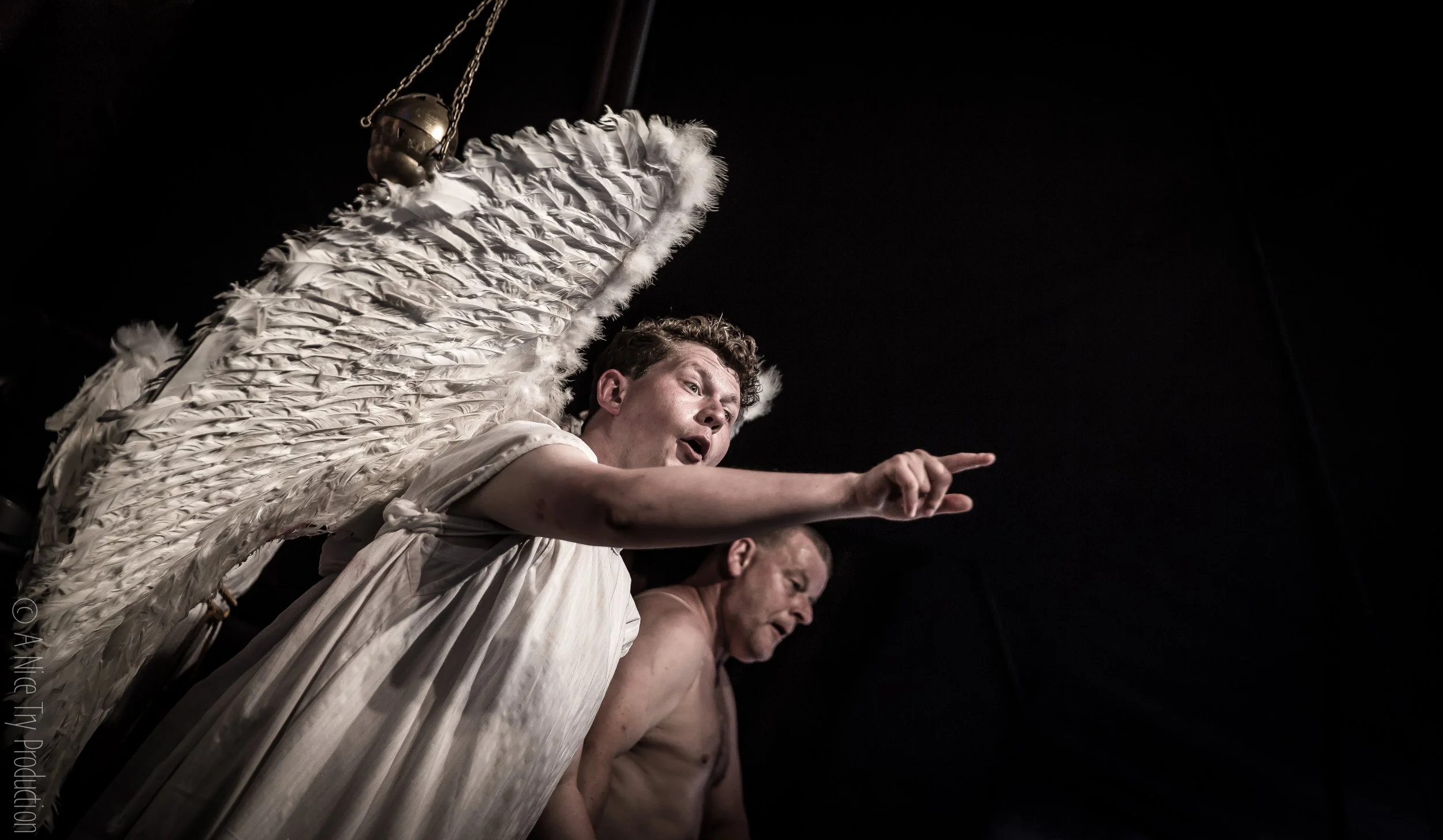 Twee acteurs in toneelkostuum, één met grote vleugels, uitdrukking van dramatische emotie, donkere achtergrond.