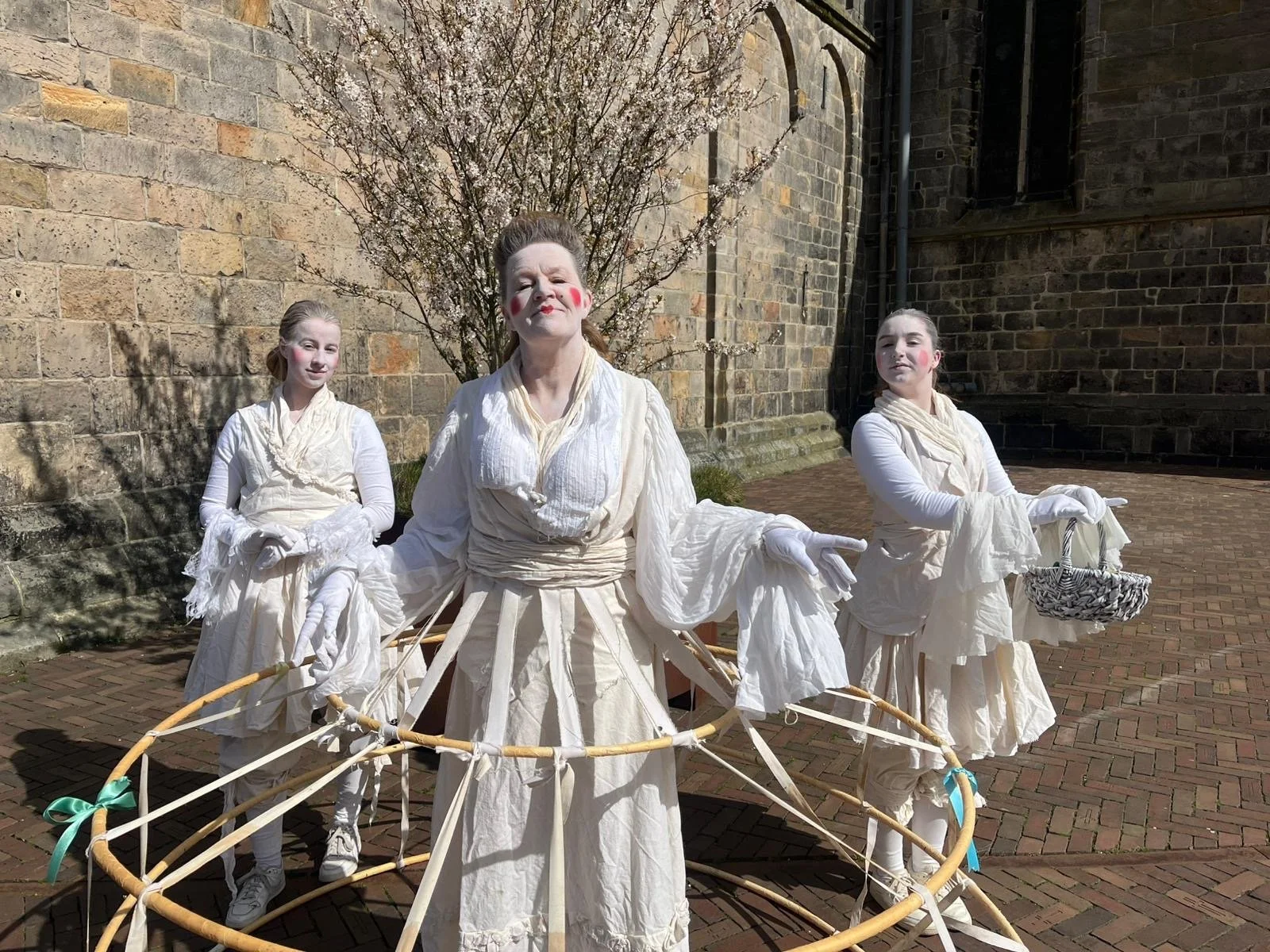 Drie mensen in witte kleding en witte handschoenen poseren buiten bij een kerk met een bloeiende boom, het lijkt op een toneelopvoering of kunstinstallatie.