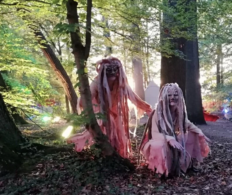 Twee grote poppen met lange, draadvormige haren en maskers, geplaatst in een bos met bomen en groen bladerdek tijdens daglicht.