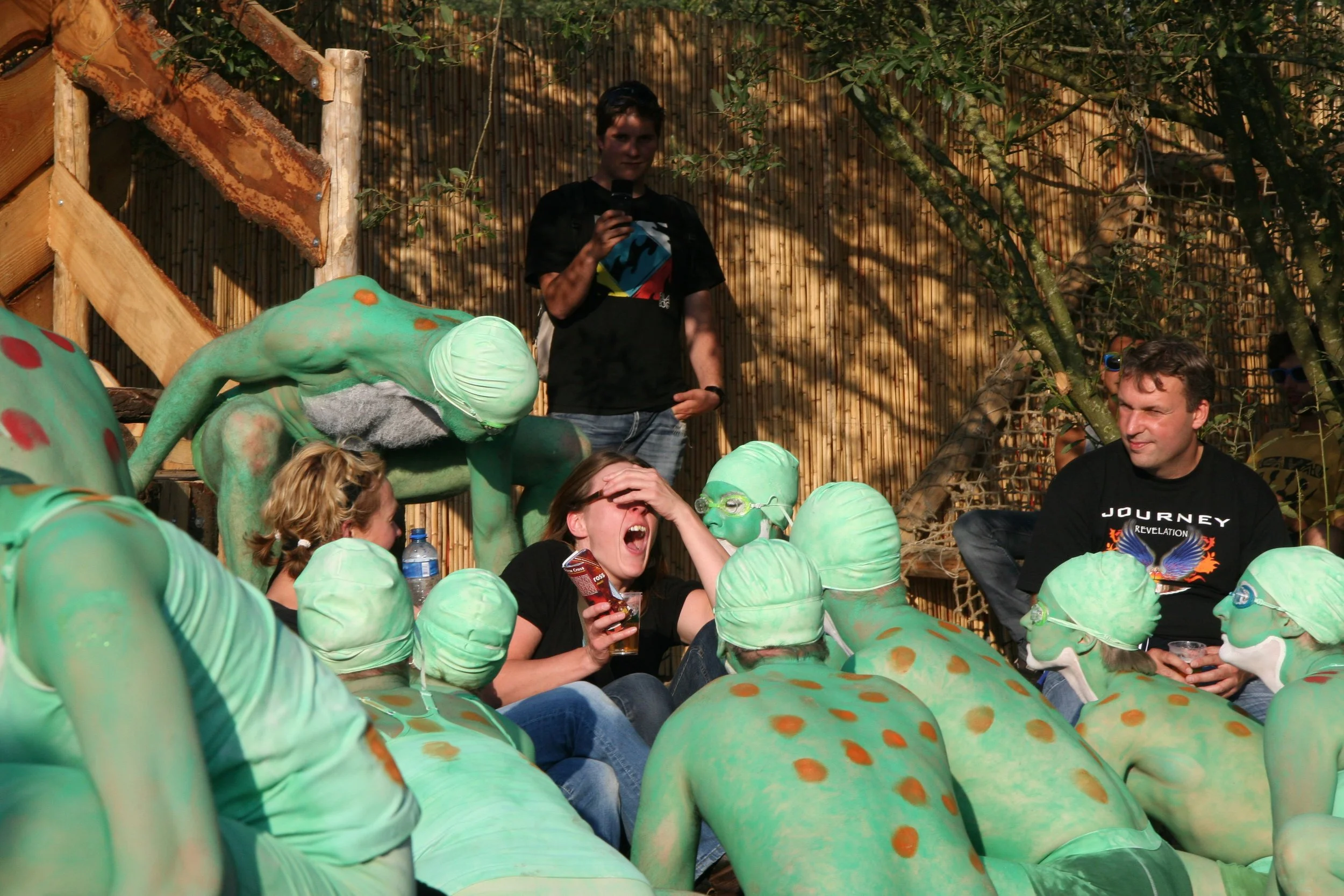 Groep mensen, inclusief enkele met groene zwemkappen en lichamen gekleed als kikkers, zitten samen in een geïmproviseerde omgeving met een houten en bamboe achtergrond. Eén persoon schreeuwt met een hand op haar gezicht.