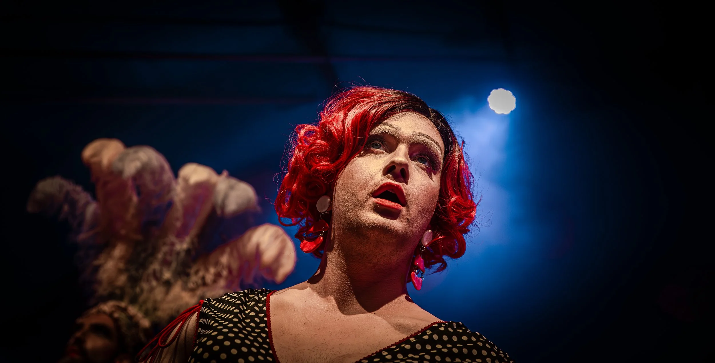 Persoon met rood gekleurd haar en opvallende oorbellen, zittend onder blauwe podiumverlichting.