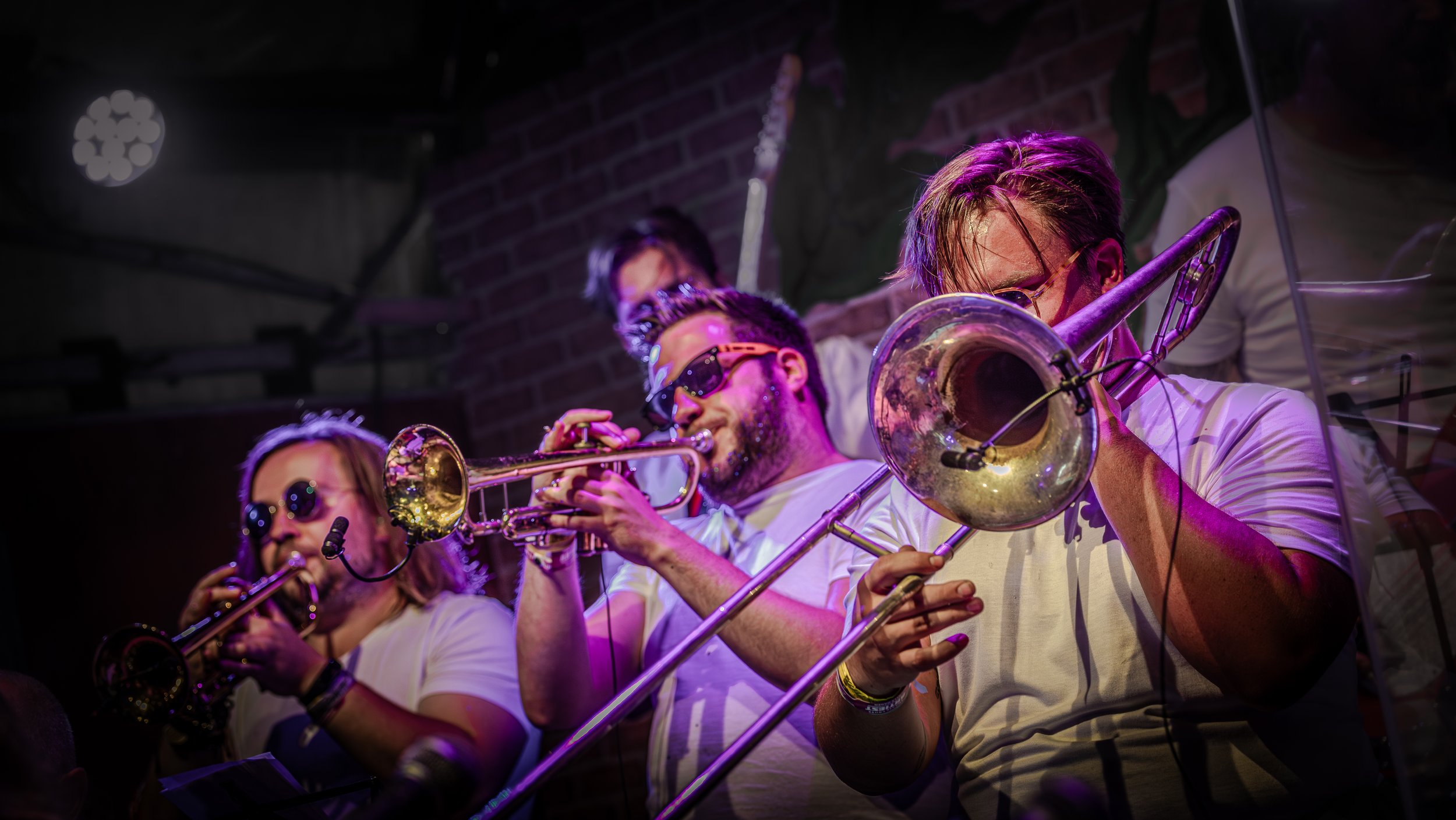Mensen die blaasinstrumenten spelen tijdens een live muziekoptreden in een club of café, met een bakstenen muur op de achtergrond en kleurrijke verlichting.