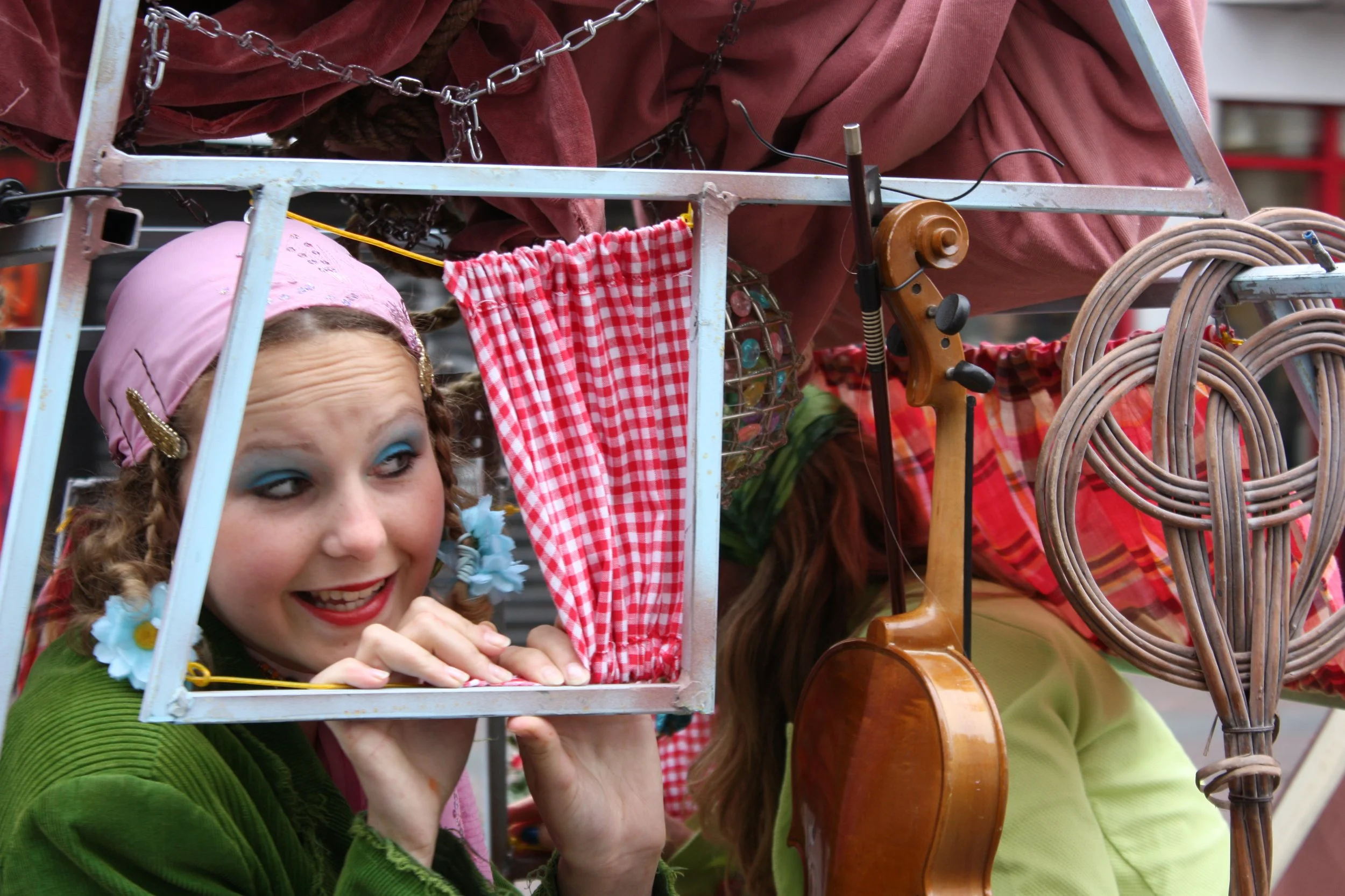 Vrouw met felgekleurde make-up en een roze hoofddoek kijkt door een raam, omringd door muziekinstrumenten en kleurrijke stoffen.