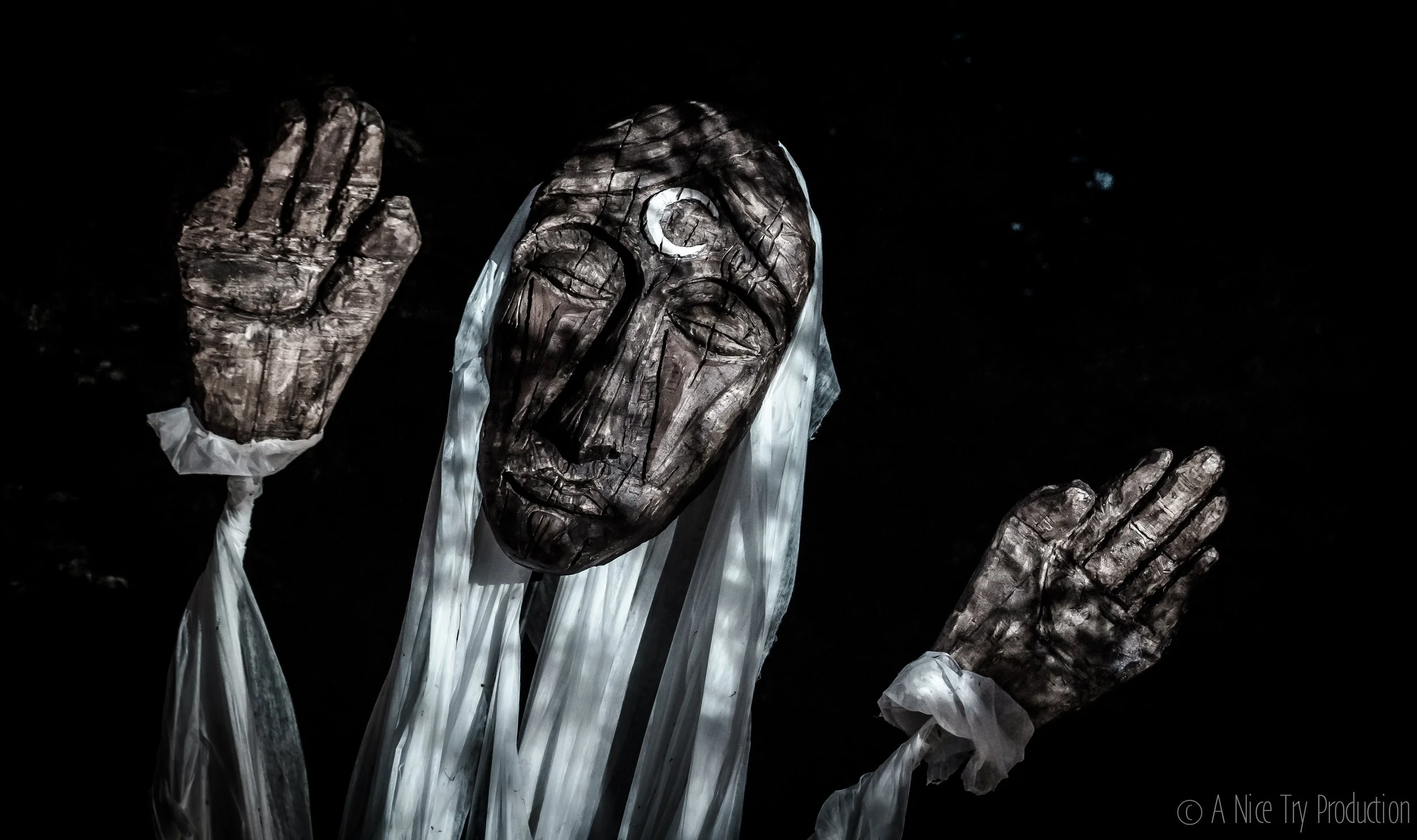 Een kunstwerk met een houten gezicht met een maan op het voorhoofd, omgeven door witte stoffen, met zwarte achtergrond.