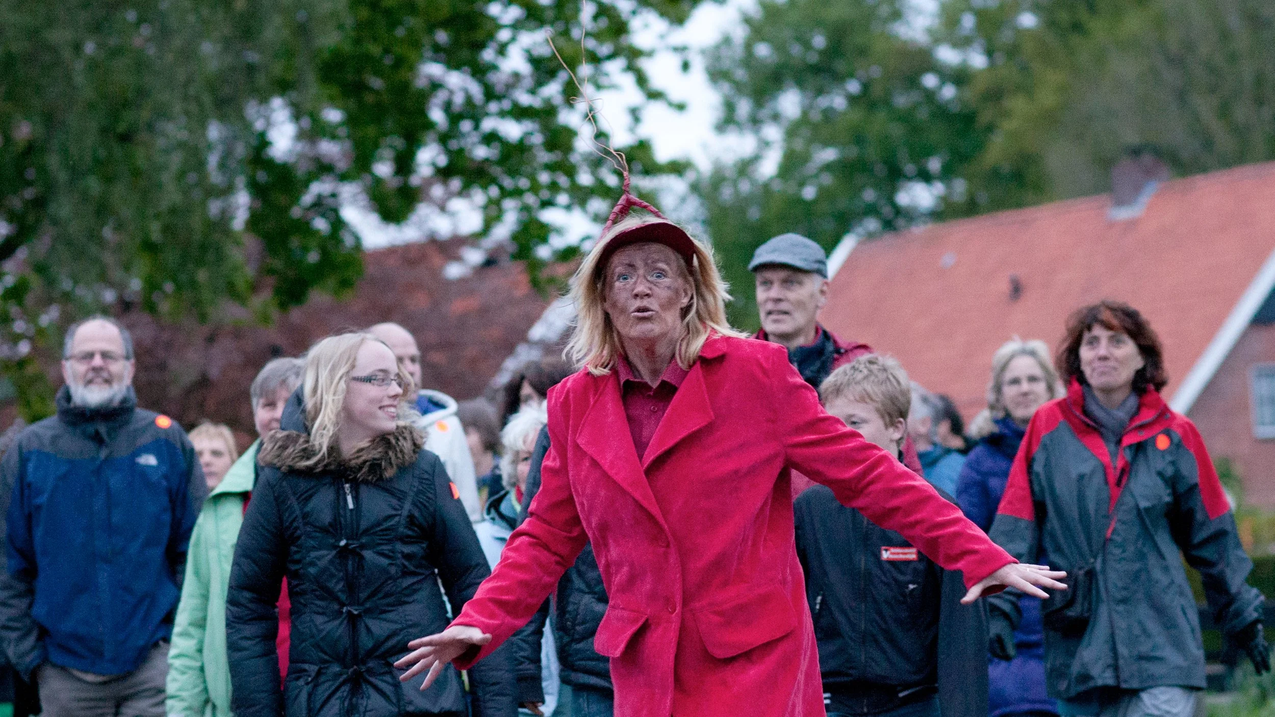 Een vrouw in een rode jas en hoed staat voor een groep mensen tijdens een buitenactiviteit.