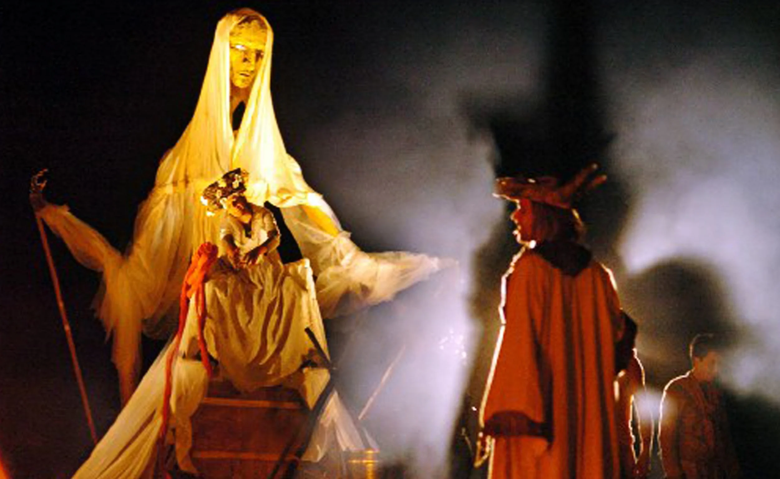 Een grote figuur in wit maskerdak, omringd door mensen in traditionele rijke kostuums, bij nacht met rook en lichteffecten.