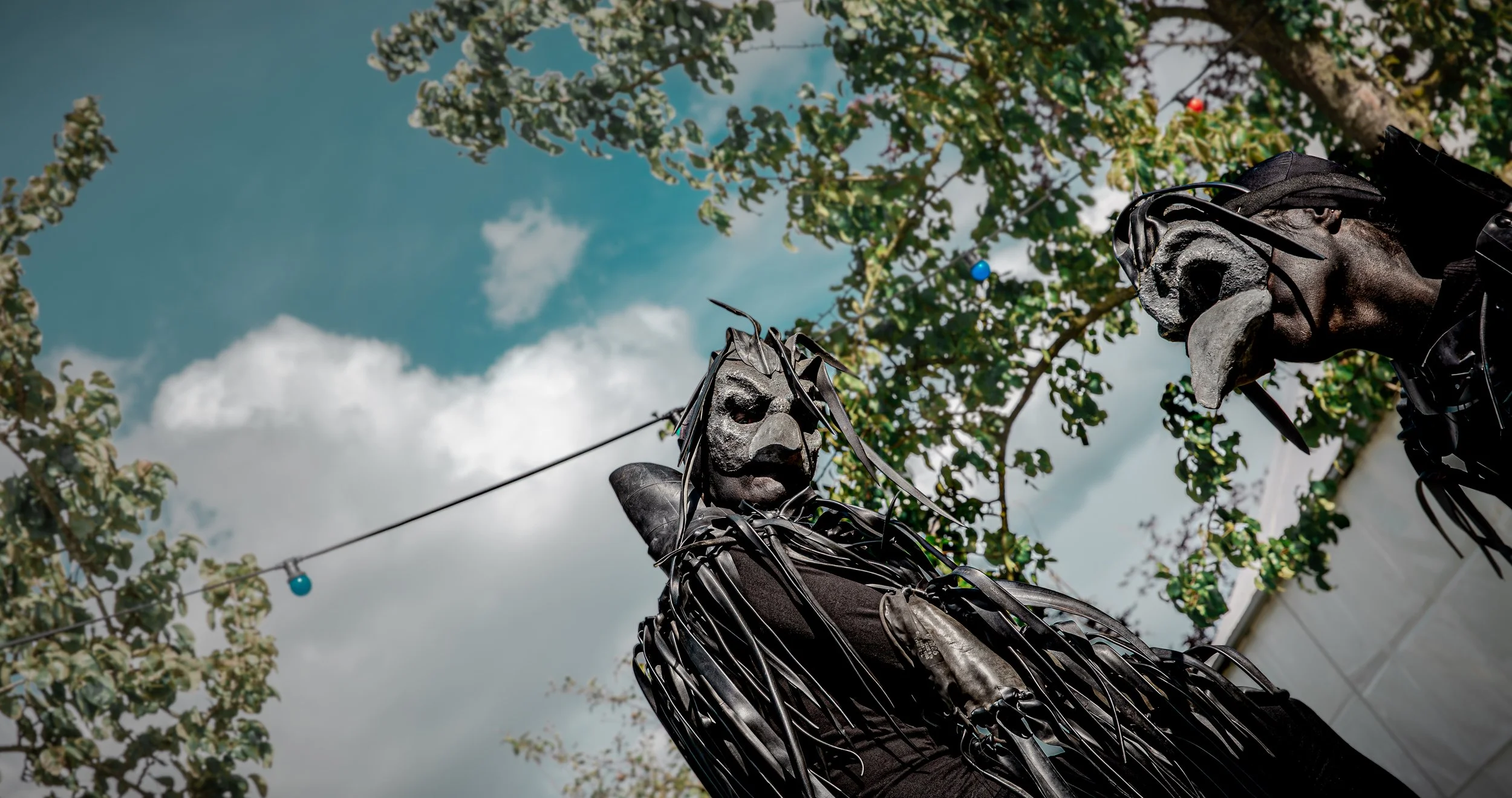 Twee mensen met ravenmaskers met een zwarte, gestructureerde huid en complexe vormen, buiten onder een blauw bewolkte lucht en groene bomen.