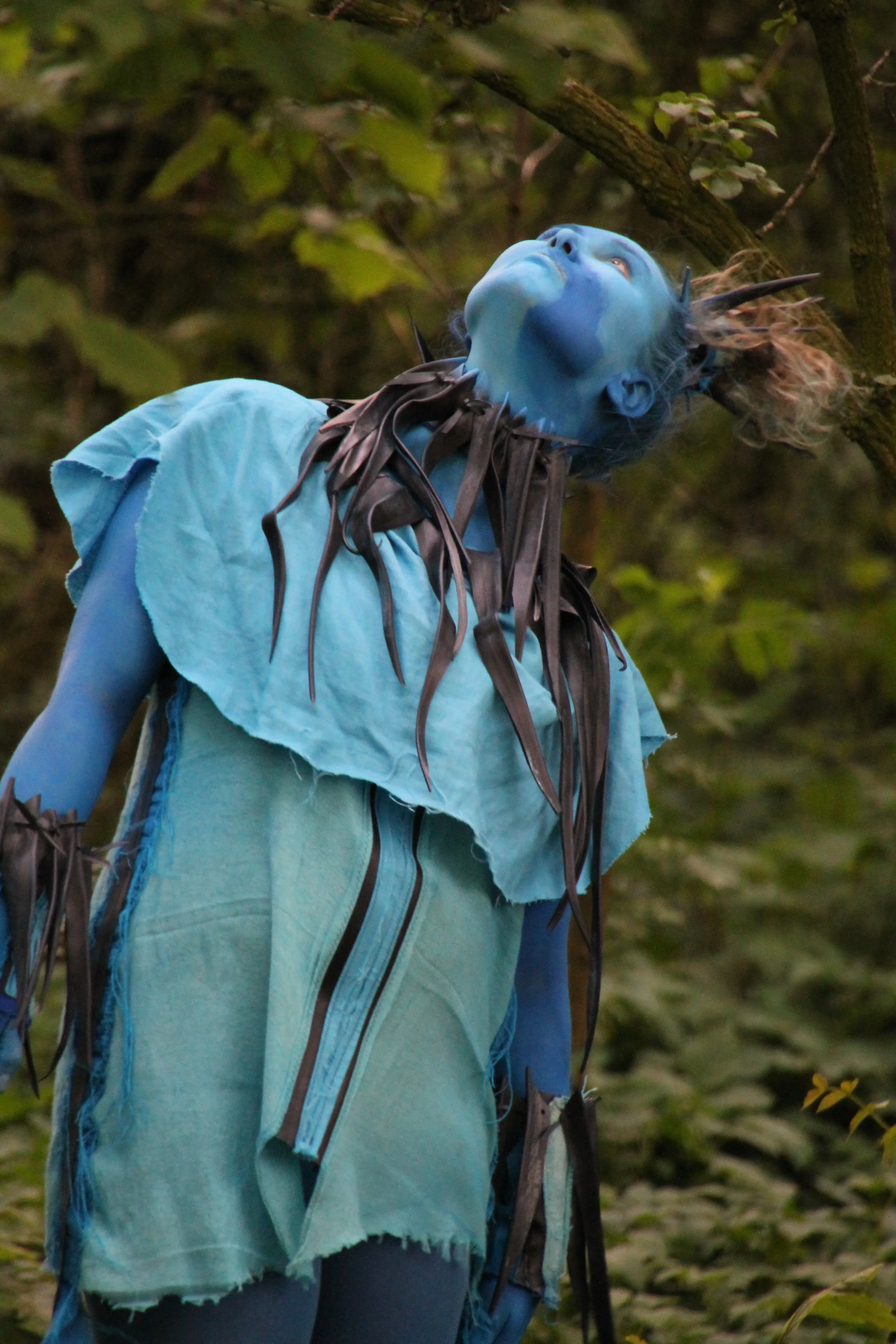 Een persoon verkleed als een blauwe naaktaard met een fantasiekostuum in een bos, met een blauwe huid en daarvan uitziende uitdrukking.
