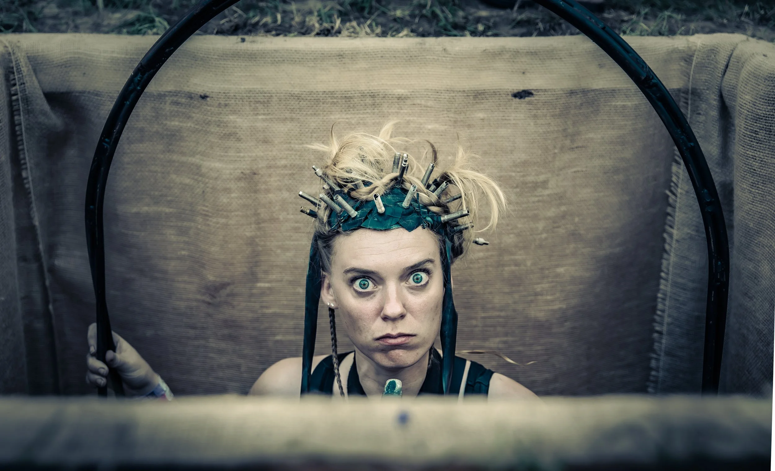 Een vrouw met felle blauwe ogen, zittend in een schommel in een kuil, kijkt recht in de camera met een ernstige uitdrukking, in een industriële setting.