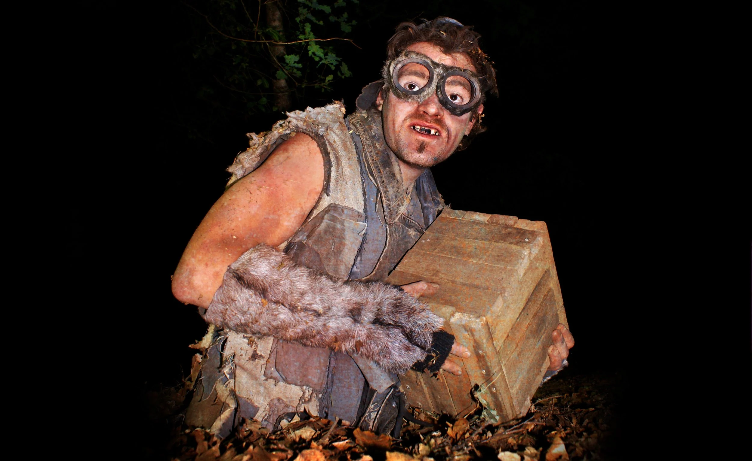 Man gekleed als een boswezen met zak, gewaad en accessoires, in een donker bos, houdend een houten kist.