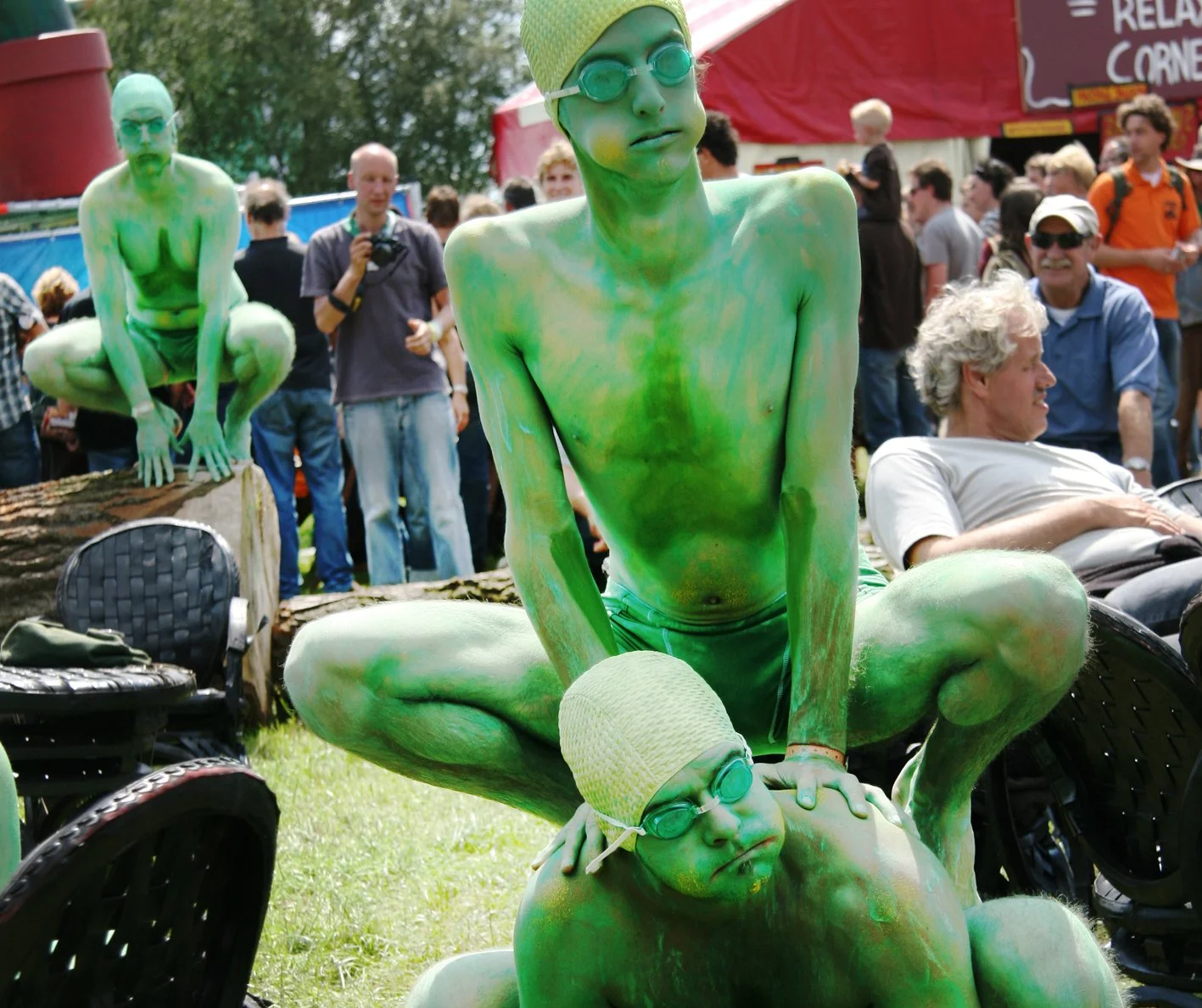 Twee mensen met volledig groene verf op hun lichaam en gezicht, een man zit op de schouders van een andere man die ook groene verf heeft. Ze dragen zwembrillen en badmutsen, op een druk festival met veel mensen op de achtergrond.