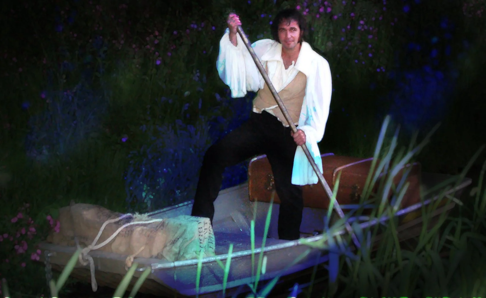 Man met een jaren 70-stijl wit shirt en zwart broek, staand in een boot met een peddel, omgeven door gras en donker blauw bloemen.