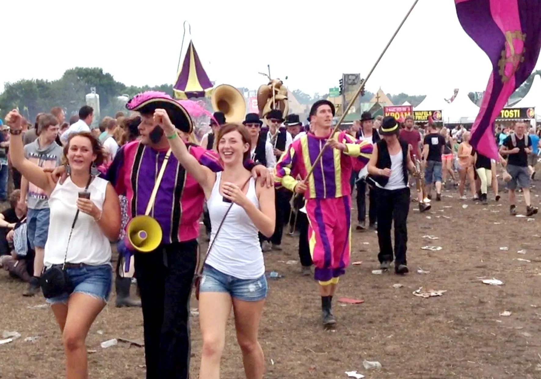 Mensen in kleurrijke kostuums vieren op een festival, met tenten en attracties op de achtergrond.