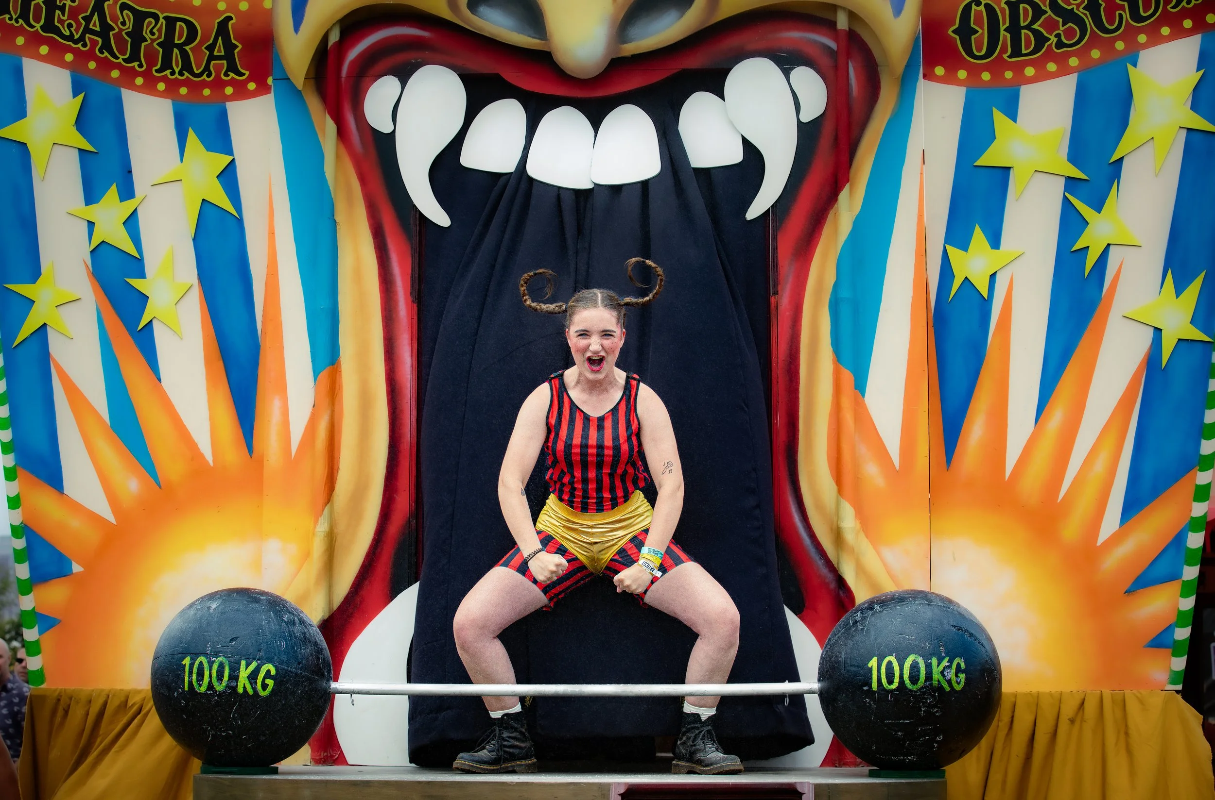 Een jonge vrouw verkleed als Pipi poseert op een circuskar, met een groot gewicht en een kleurrijke achtergrond met sterren en explosies.