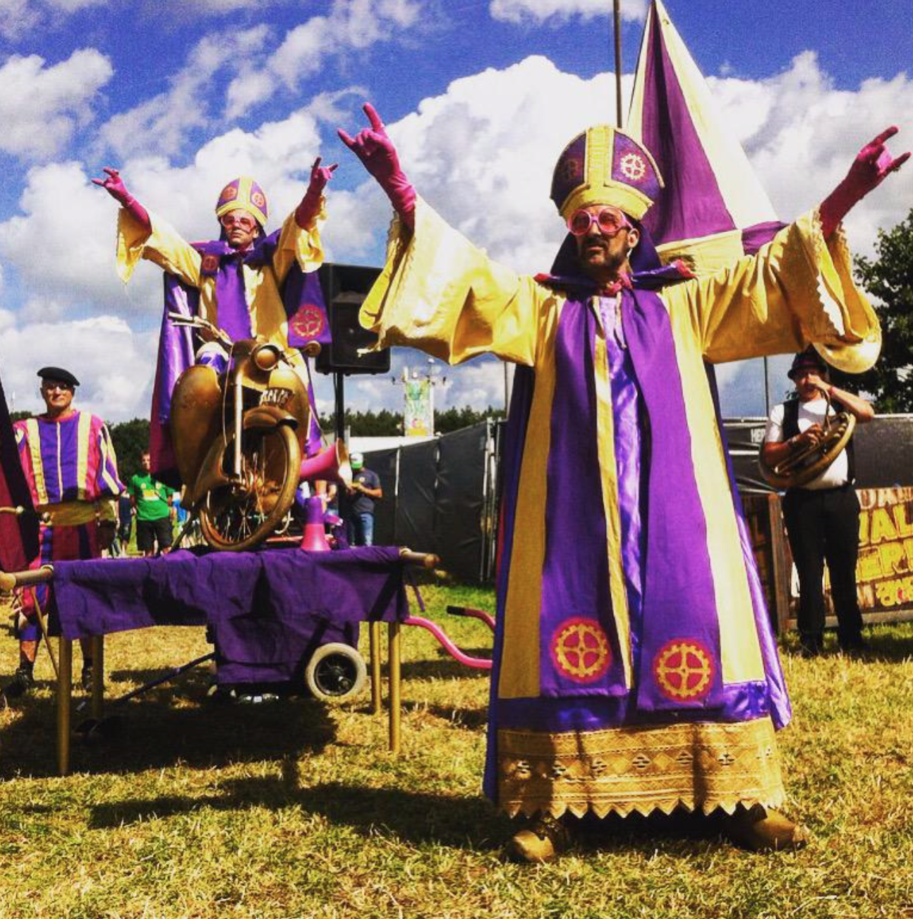 Mensen in kleurrijke, carnavaleske kostuums met paarse en gele kleuren, dansend en spelend tijdens een festival, met een achtergrond van een blauwe lucht met wolken en groene bomen.