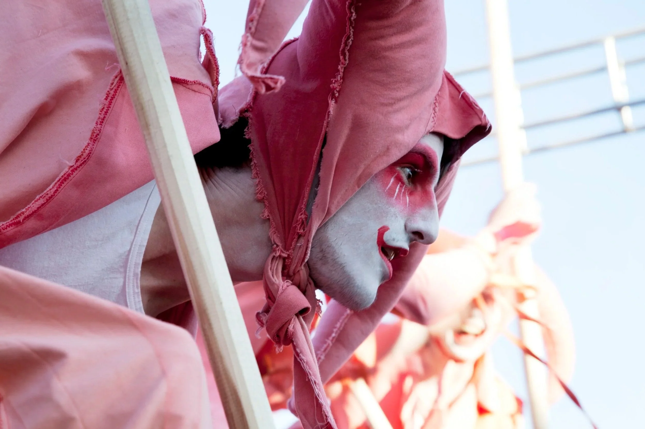 Close-up van een nar in een roze Kostuum met gezichtsmake-up en een puntige hoed, met andere vergelijkbare figuren op de achtergrond.