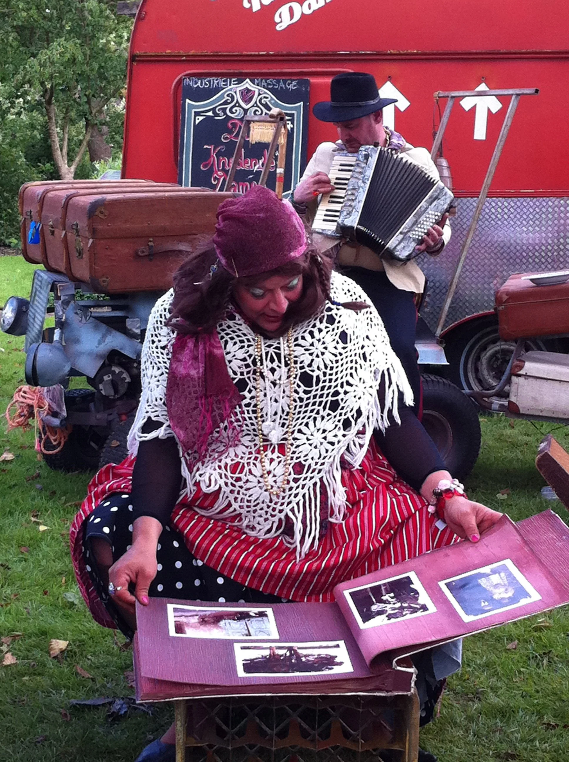 Een vrouw die op een grasveld zit en in een album met foto's kijkt, terwijl een man op een harmonica speelt op de achtergrond.