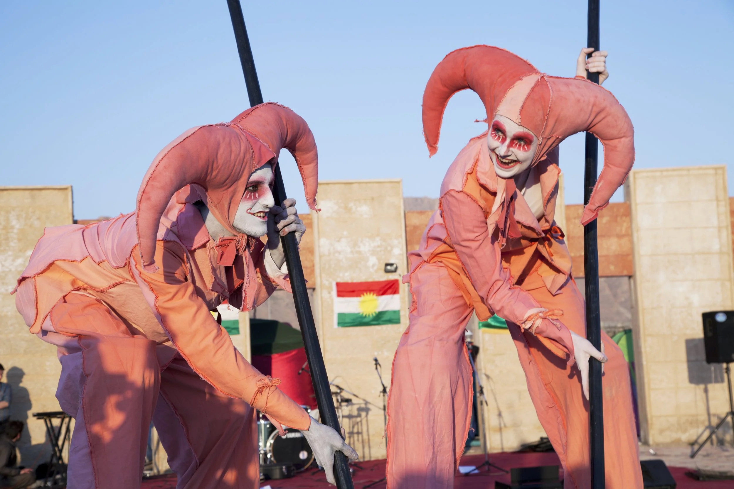 Twee narren met roze kostuums en hoedachtige stukken op hun hoofden, danst op een podium met een achtergrond van het Kurdistan-vlag en een stenen muur.