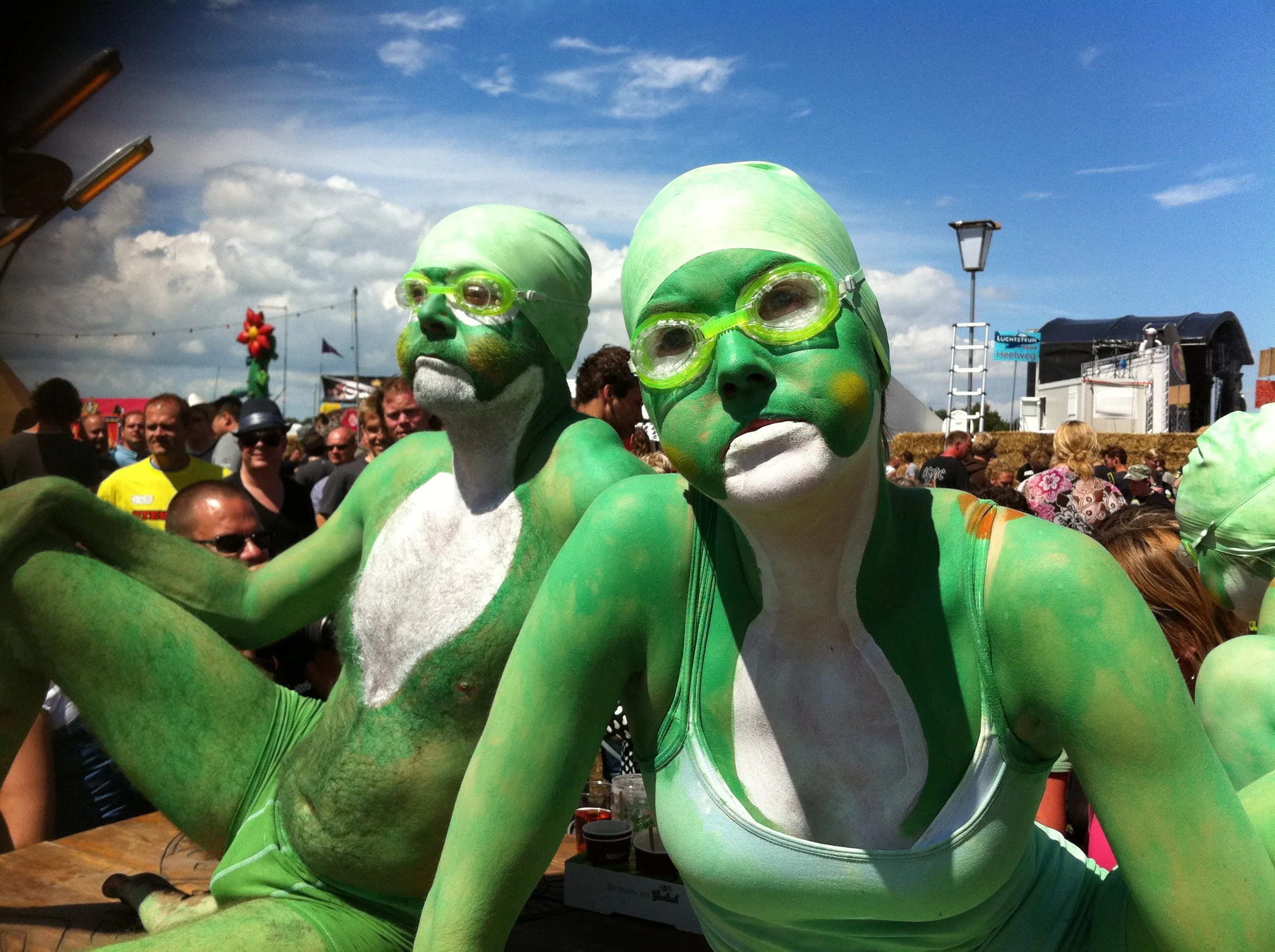 Twee mensen in groene lichaamsverf en zwembrillen poseren op een festival, met een menigte en kraampjes op de achtergrond onder een blauwe lucht met wolken.