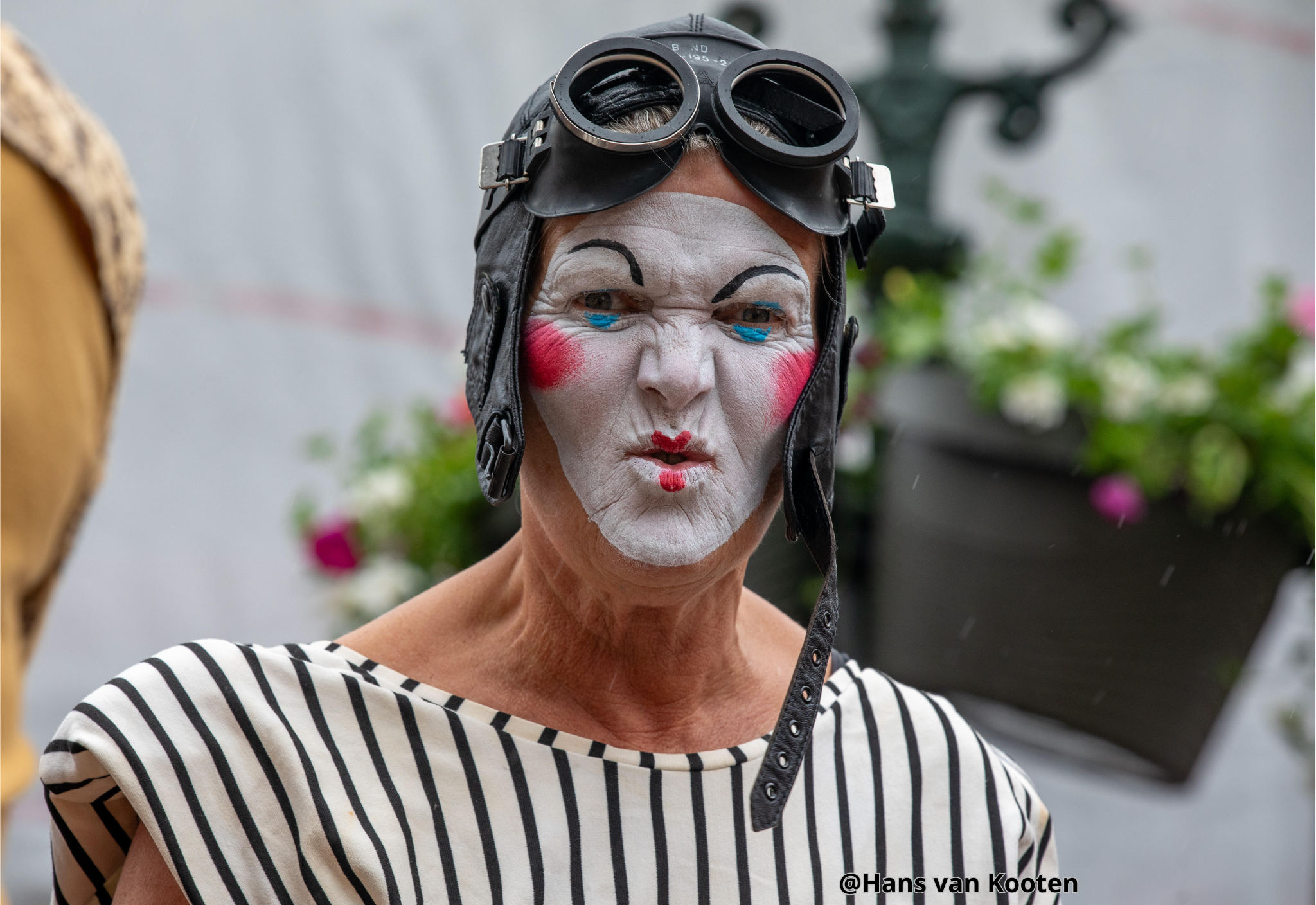 Een oudere vrouw met clownsmake-up, een zwart leren helm met bril, en een zwart-wit gestreepte blouse.
