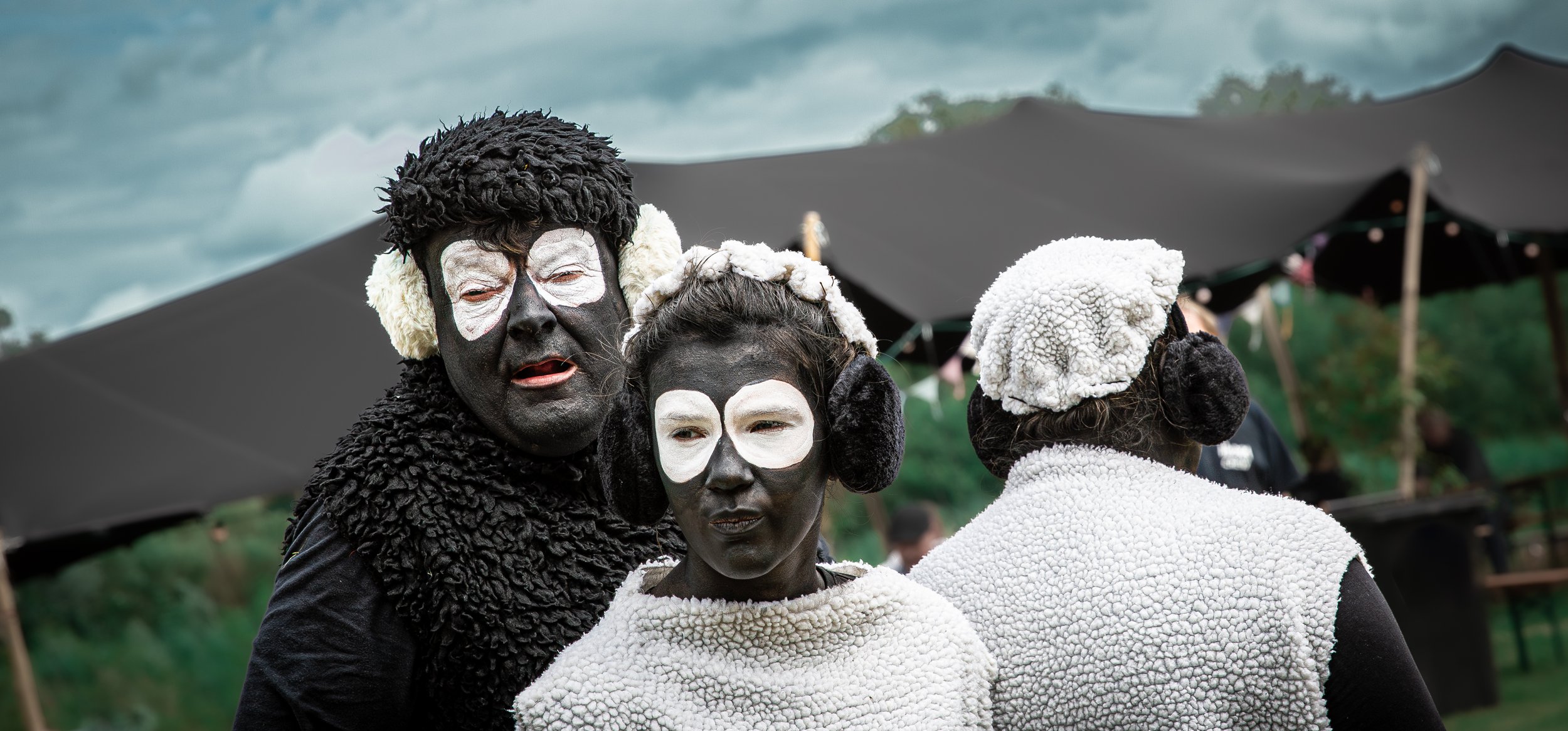 Drie mensen in zwarte en witte schapenkostuums met gezichten geschminkt als schapen, voor een zwart tentje, buiten.
