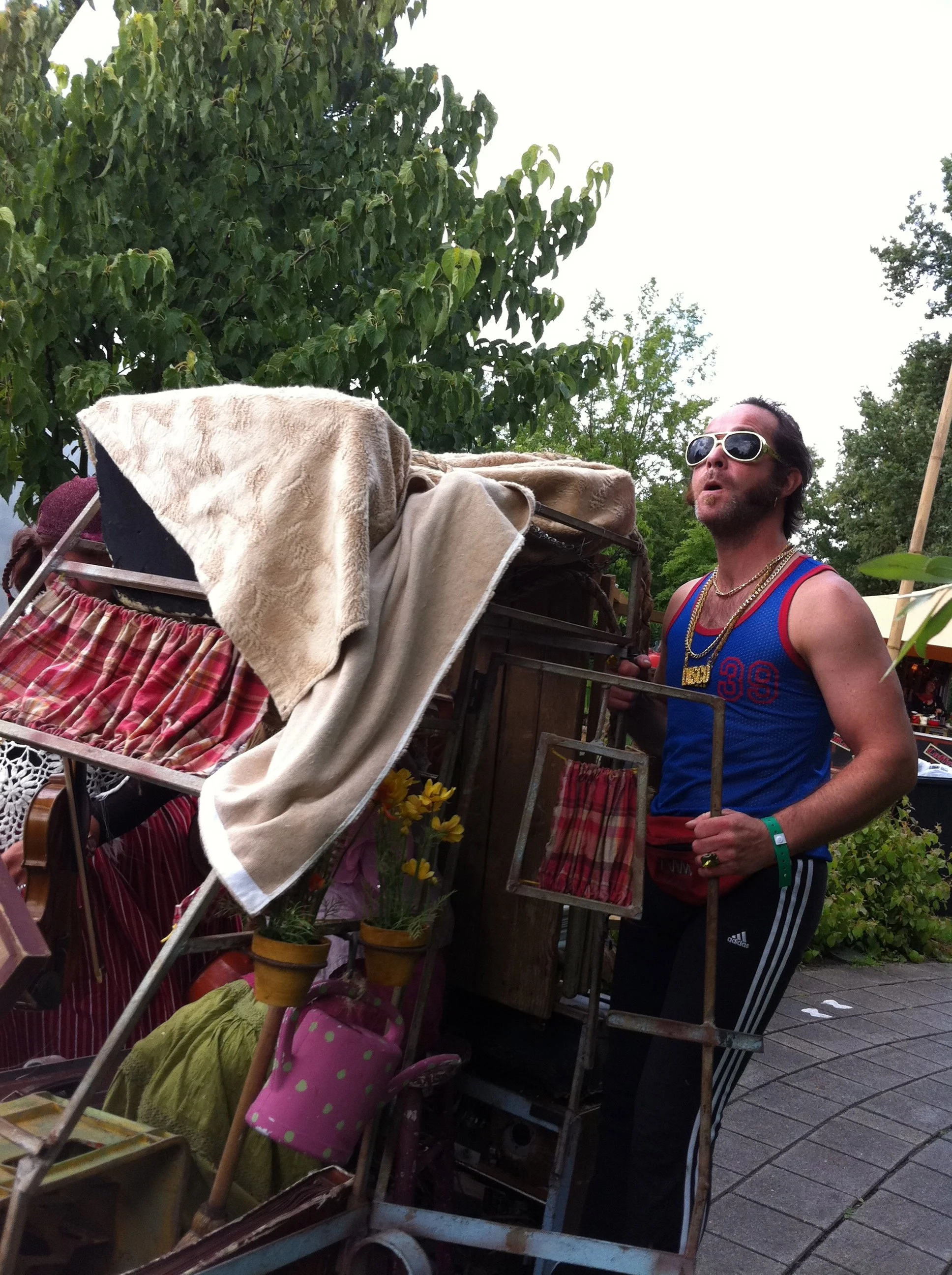 Man met donkere haar, zonnebril en kettingen, in sportoutfit, staat naast een met spullen geladen kar op een markt, omringd door groen.