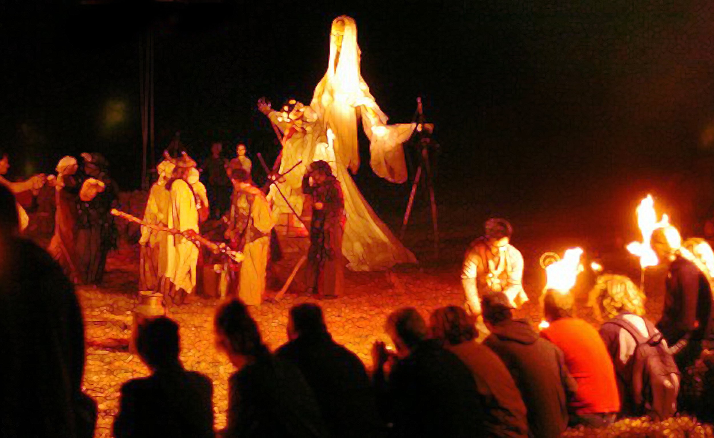 Theater performance of a religious scene with a large glowing statue and actors around fire at night, audience watching.