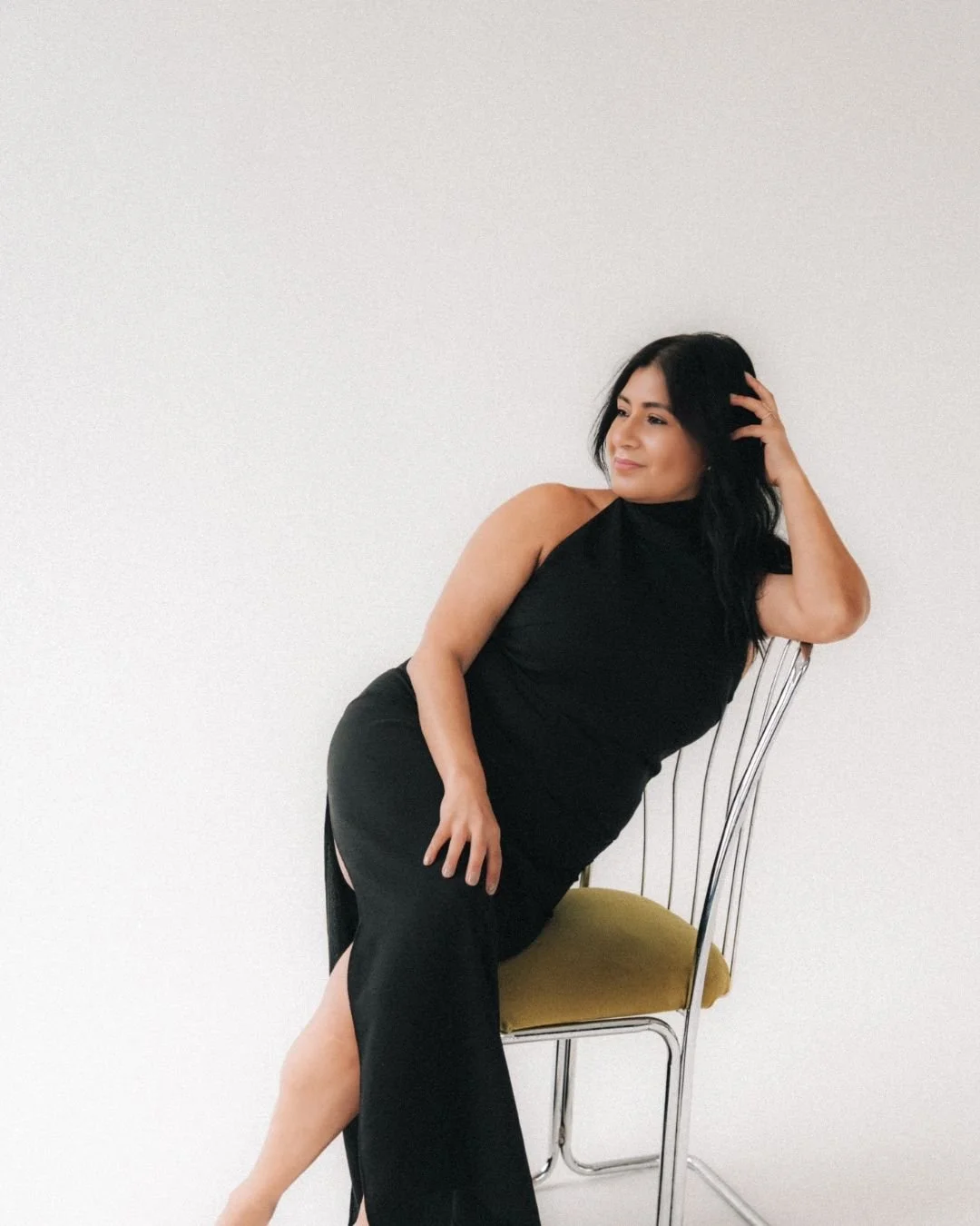 A woman with black hair and a black dress sitting on a metal chair with a yellow cushion, posing against a plain white wall.
